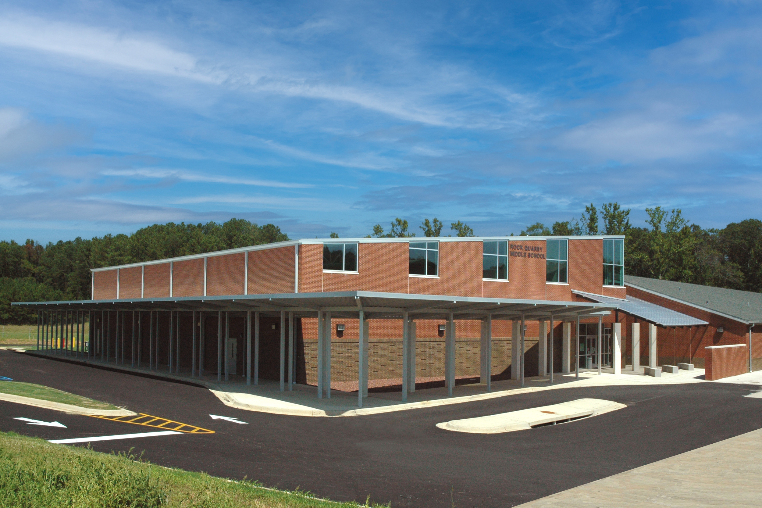 New brick middle school building with large windows, a covered walkway, and a parking lot in front, surrounded by trees and a blue sky.