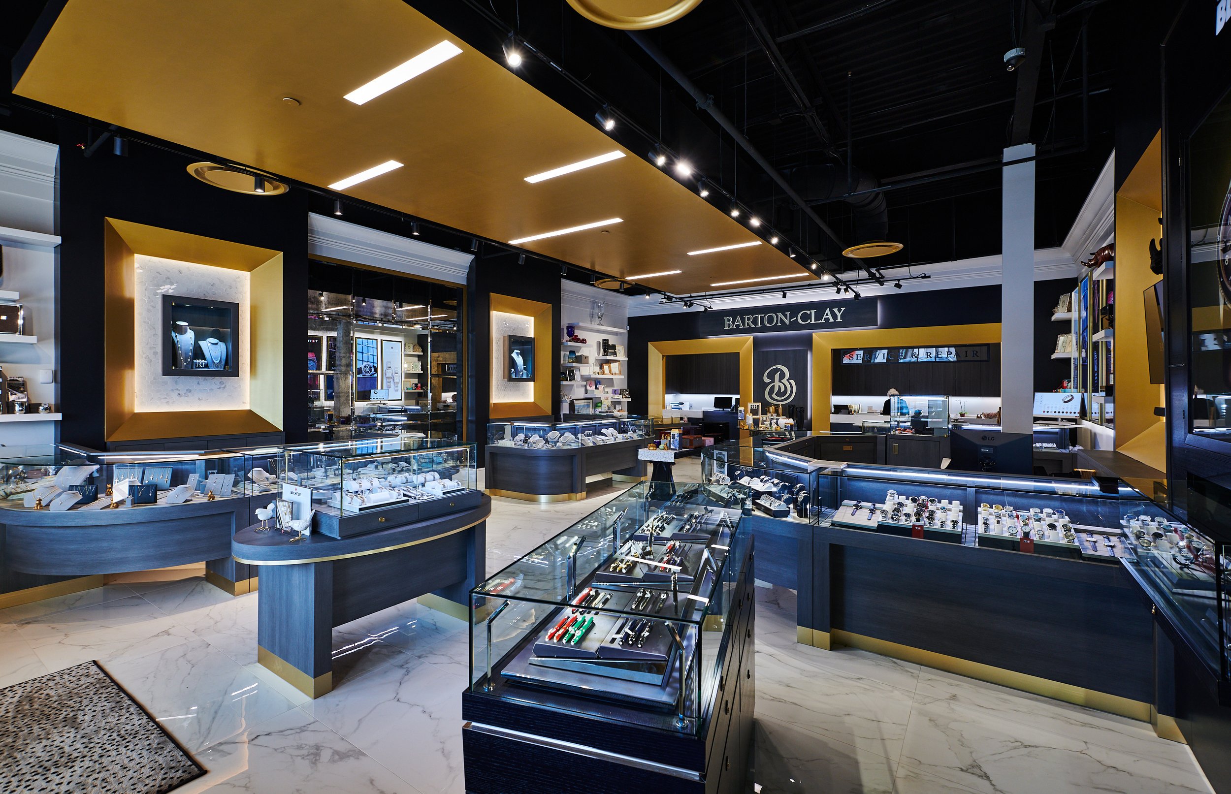 Interior of Barton Clay Fine Jewelers with display cases, shelves of jewelry, and a gold ceiling cloud. A sign reads 'Barton-Clay' at the back. project by B Group Architecture, Inc 
