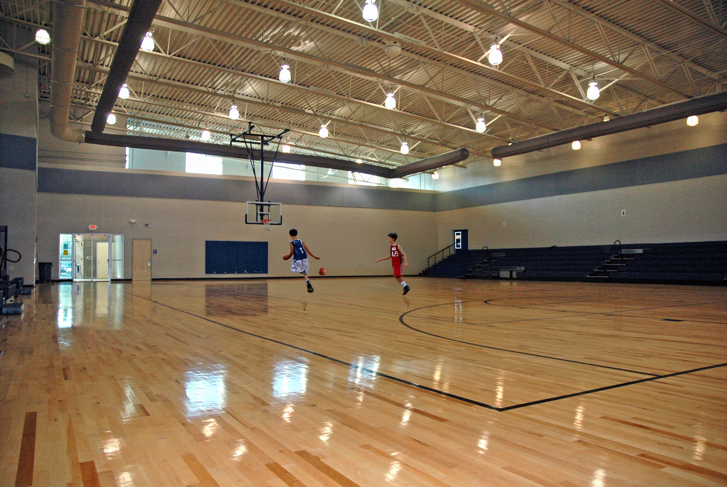 An indoor basketball court with two children playing, one dribbling a basketball in a blue jersey, and the other in a red jersey. The court has a polished wooden floor, high ceilings with multiple lights, and seating on the right side.
