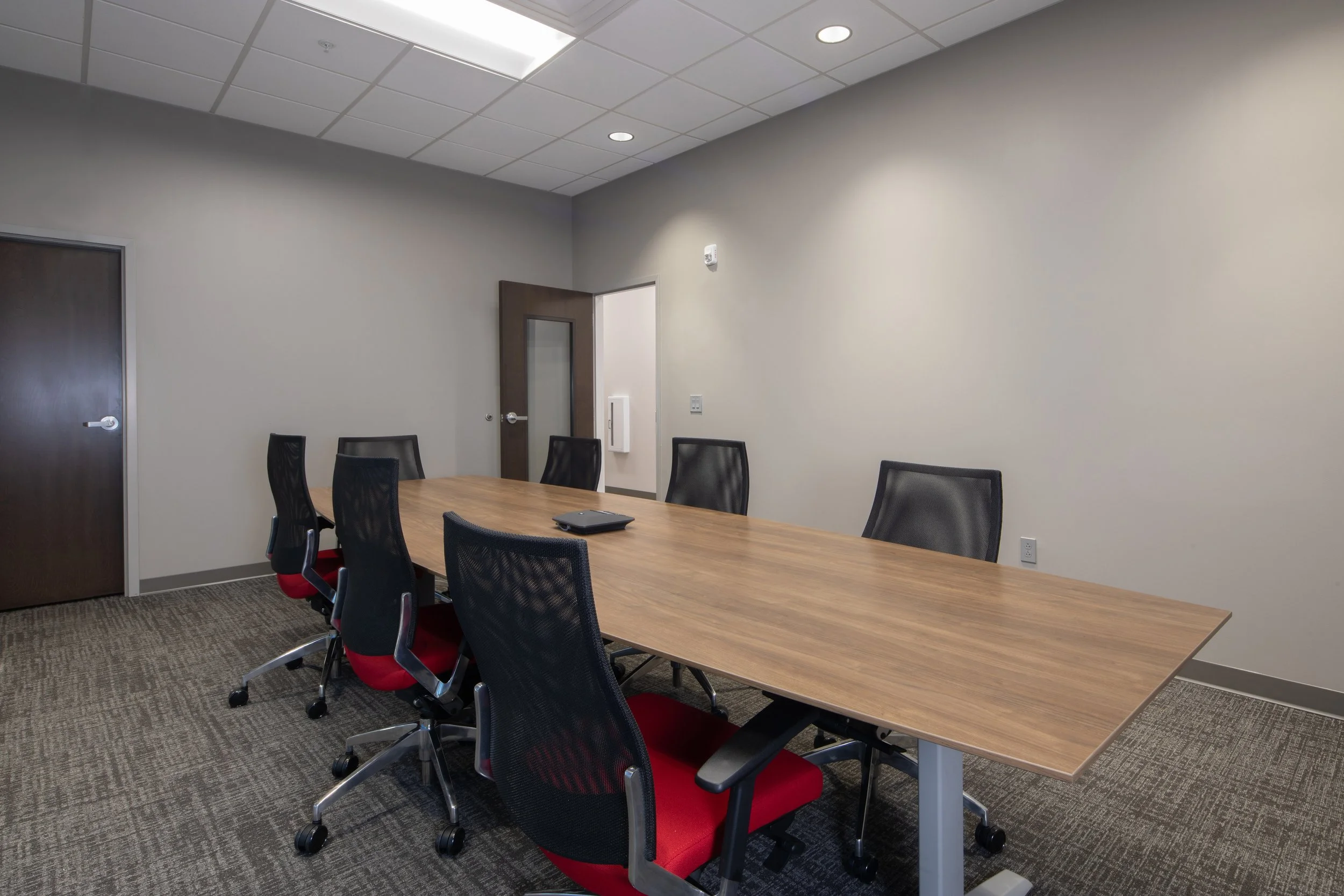 Conference room with a long wooden table, six black chairs with red seats, and two closed doors in a modern office setting.