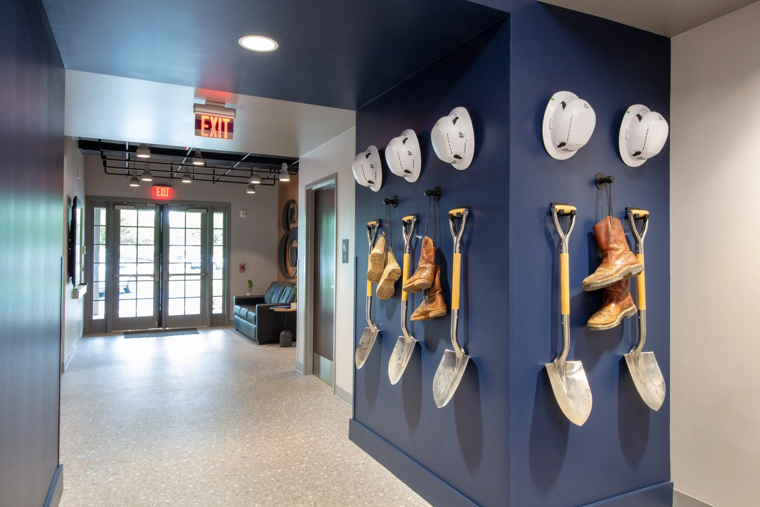 Interior of a modern building with a blue wall displaying five white helmets, five shovels with boots hanging from chains, and an exit sign. In the background, a seating area and glass entrance doors are visible.