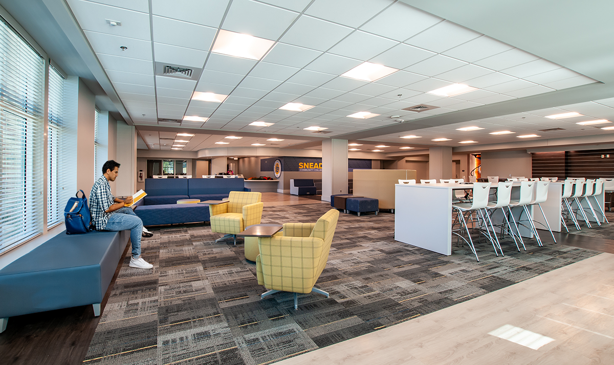 A spacious indoor lounge with large windows, seating areas with chairs and sofas, and a high ceiling with grid lights.