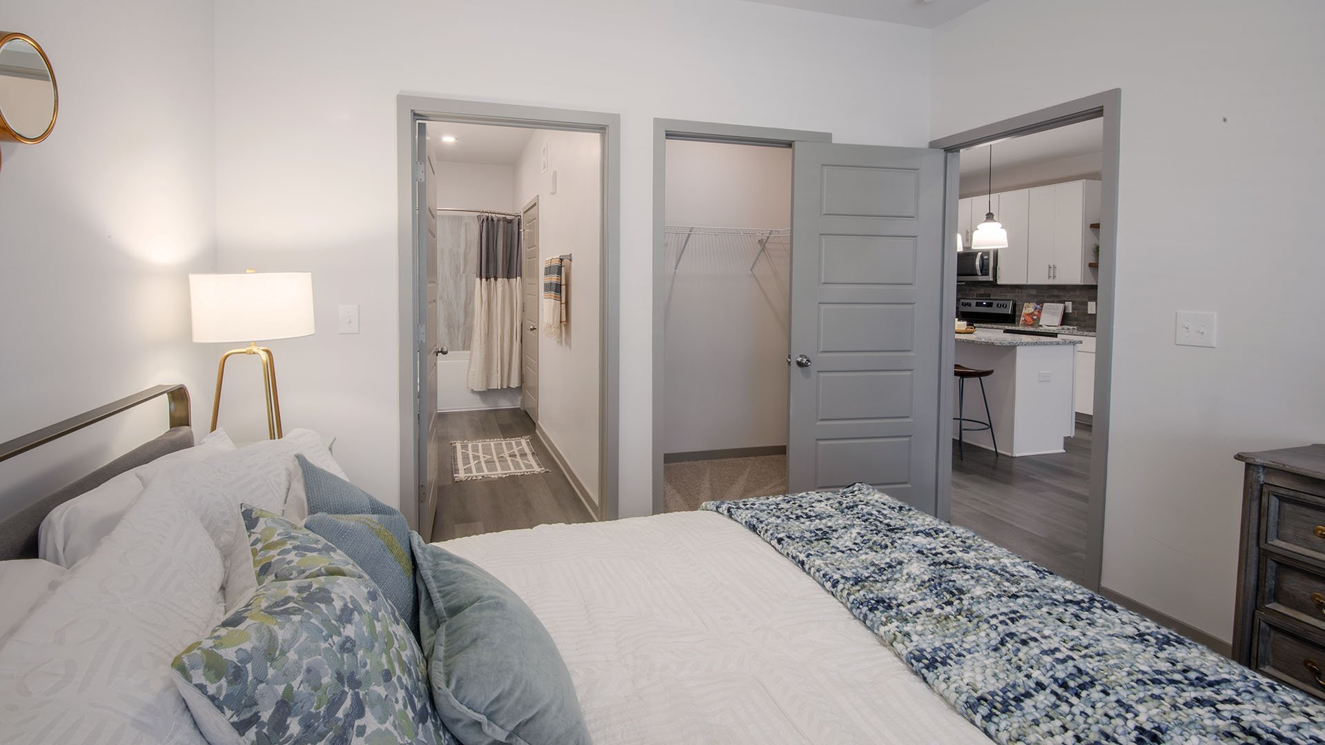 One bedroom in Axel Row Apartments with bed, pillows, bedside lamp, view into bathroom, walk-in closet, and kitchen in the background. - project by B Group Architecture, Inc.