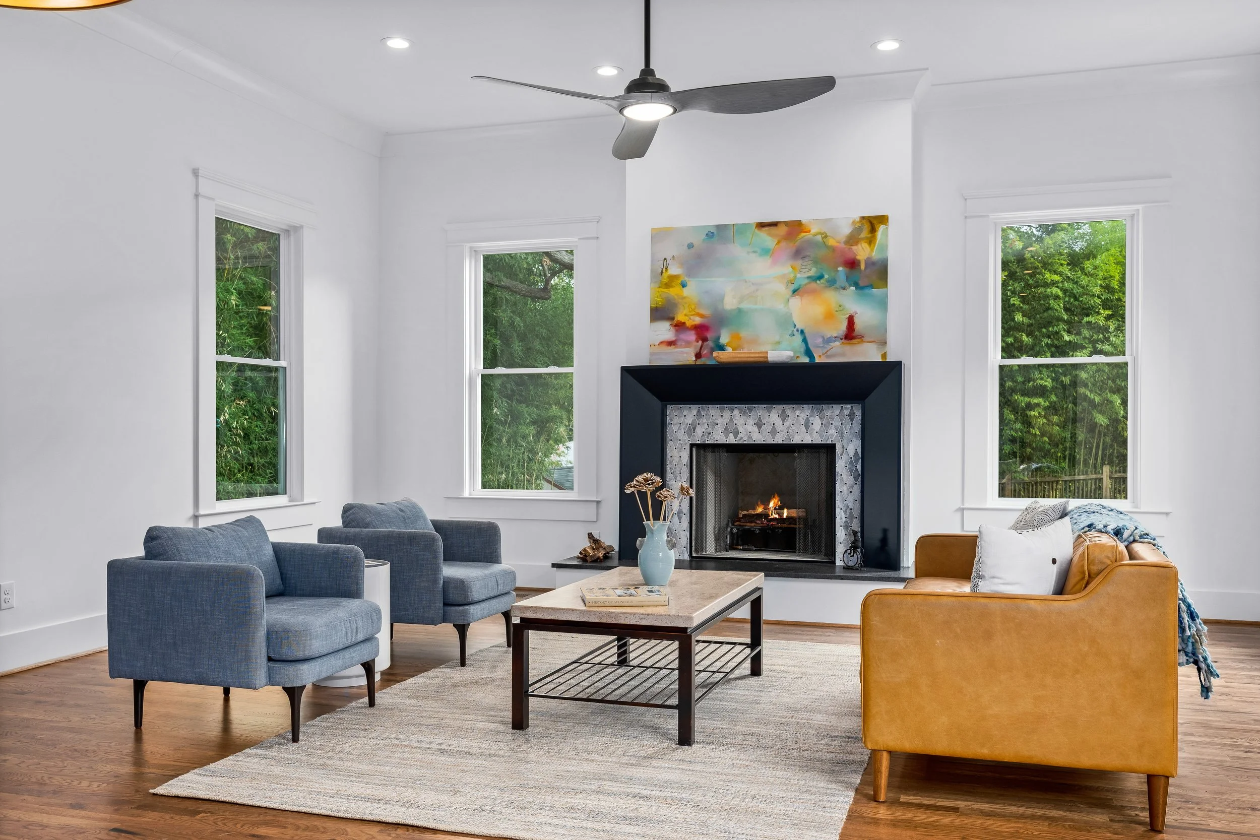 Living room with a white wall, three windows allowing natural light to shine in. The historic fireplace was renovated with tile and a colorful abstract painting above, a ceiling fan, and furniture including two blue armchairs and a mustard-colored so