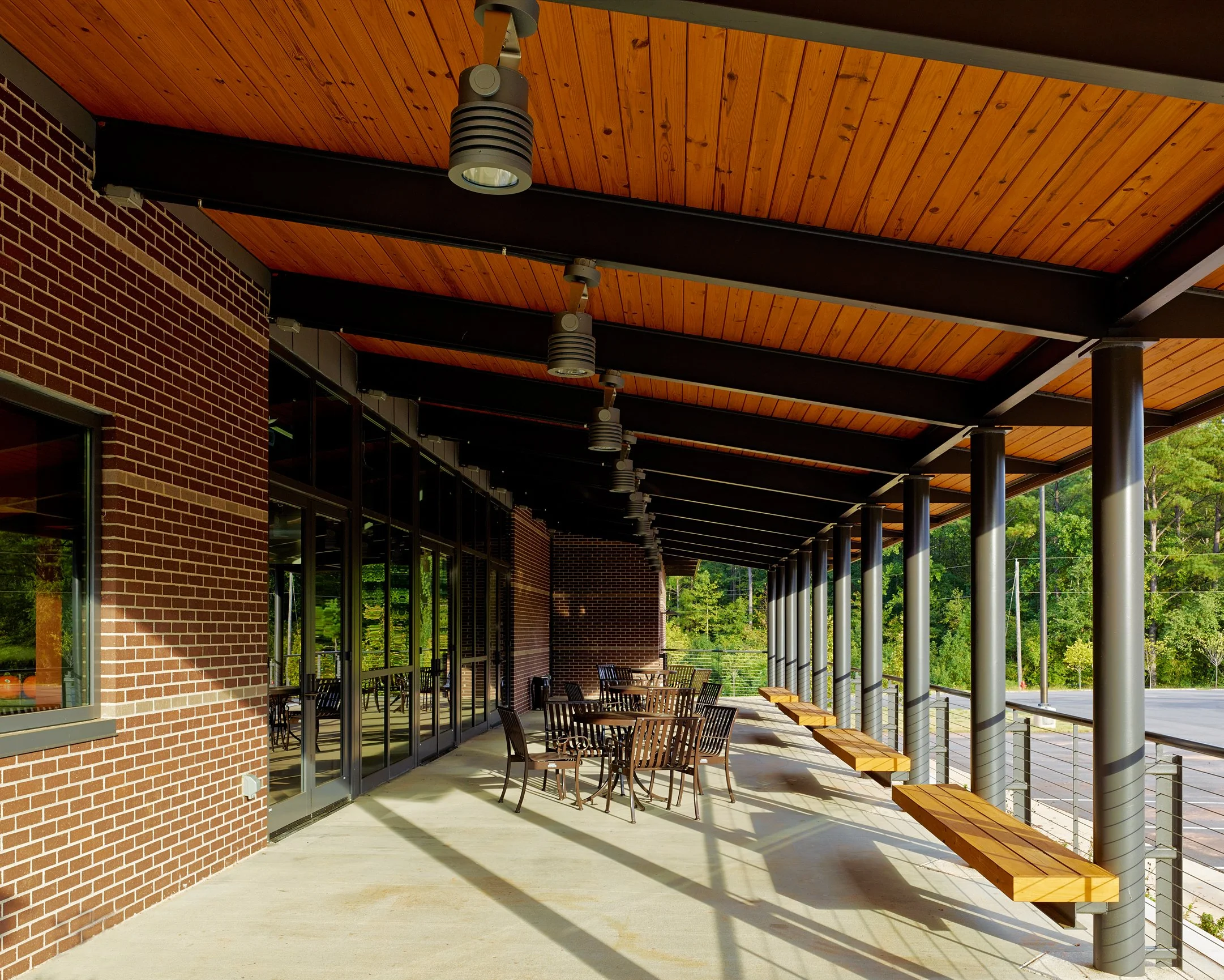 Outdoor patio with brick wall, glass windows, wooden ceiling, and metal and wooden benches, surrounded by green trees.