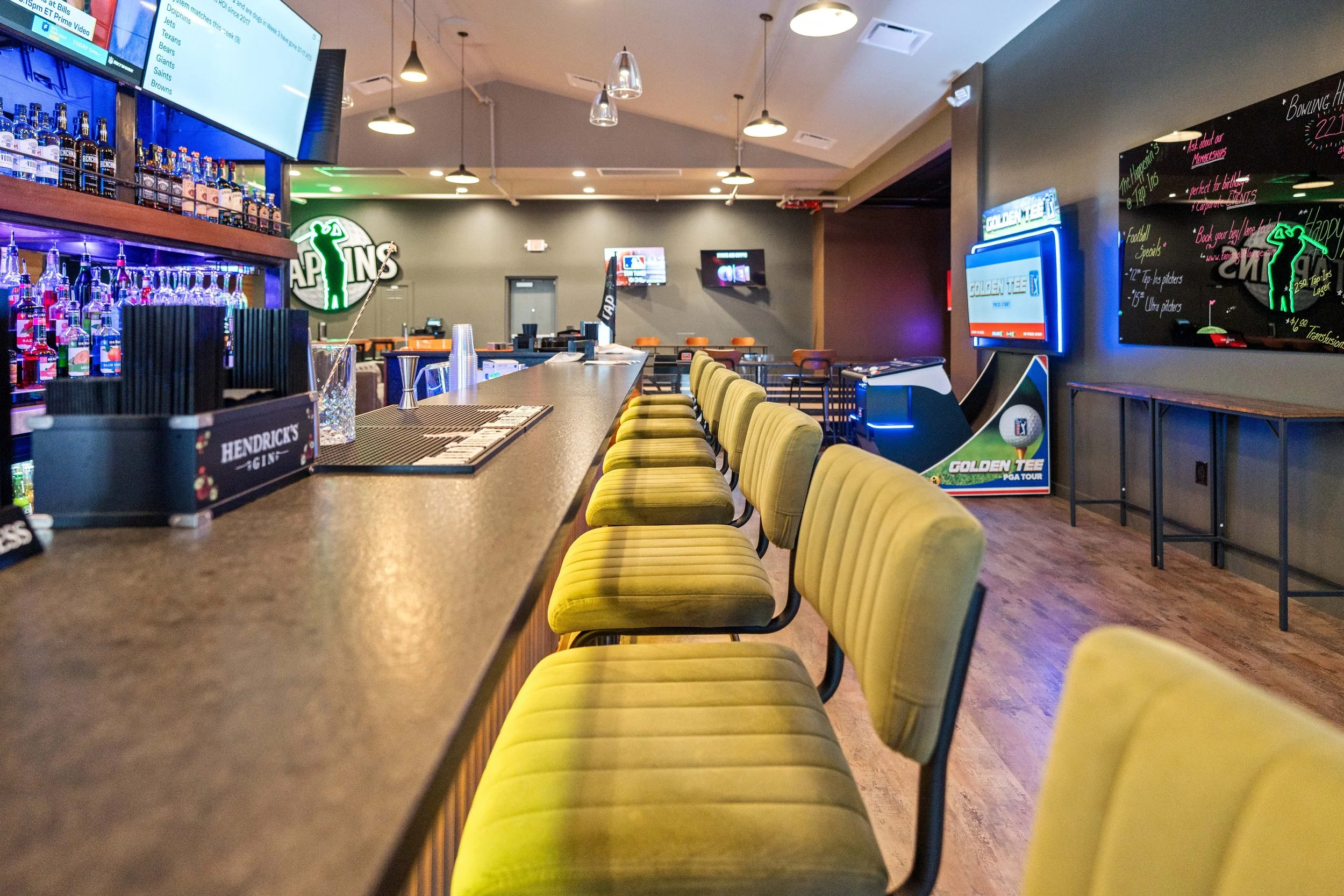 Tap Ins Bar with yellow upholstered bar seats  lined up along the honed granite counter, neon signs, TV screens, and some gaming machines in the background. project by B Group Architecture, Inc