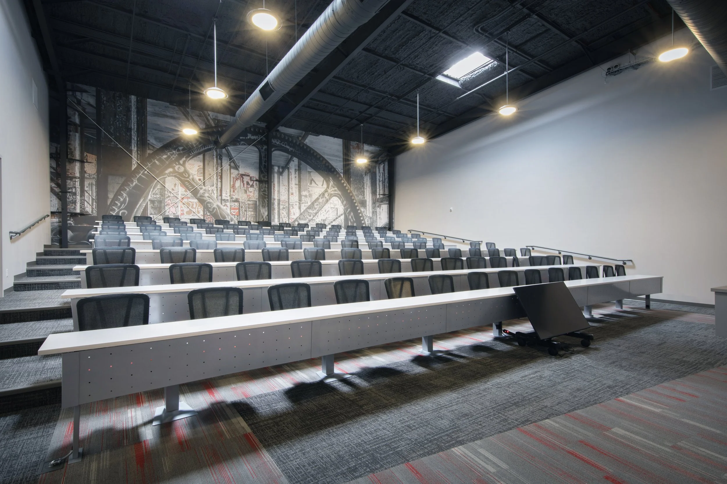 Motion Industries lecture hall with tiered seating, black mesh chairs, white desks, gray carpeting, industrial ceiling with air ducts and hanging lights, custom graffiti mural on the back wall.  project by B Group Architecture, Inc. 