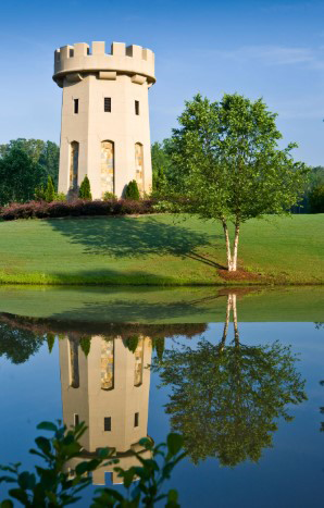 Ballantrae Golf Club stone tower with battlements stands on a grassy hill next to a small lake. A green tree and shrubs are also visible, with their reflection in the water. The sky is clear and blue. project by B Group Architecture Inc. 