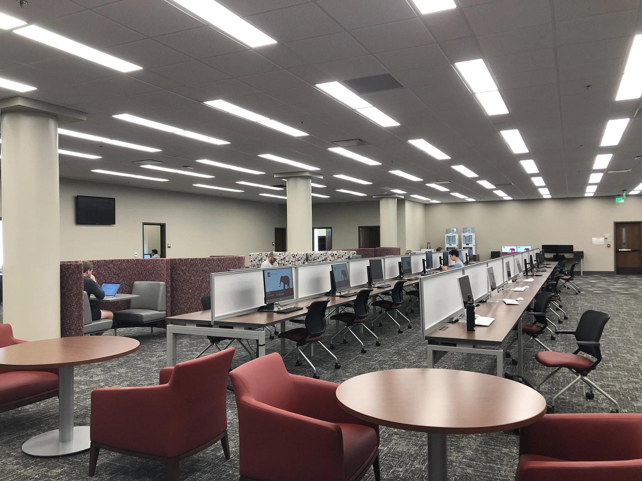 A spacious library with multiple rows of workstations.  The room features a high ceiling with linear LED lighting, and a mix of lounge and desk seating, including round tables with red chairs.. - project by B Group Architecture, Inc
