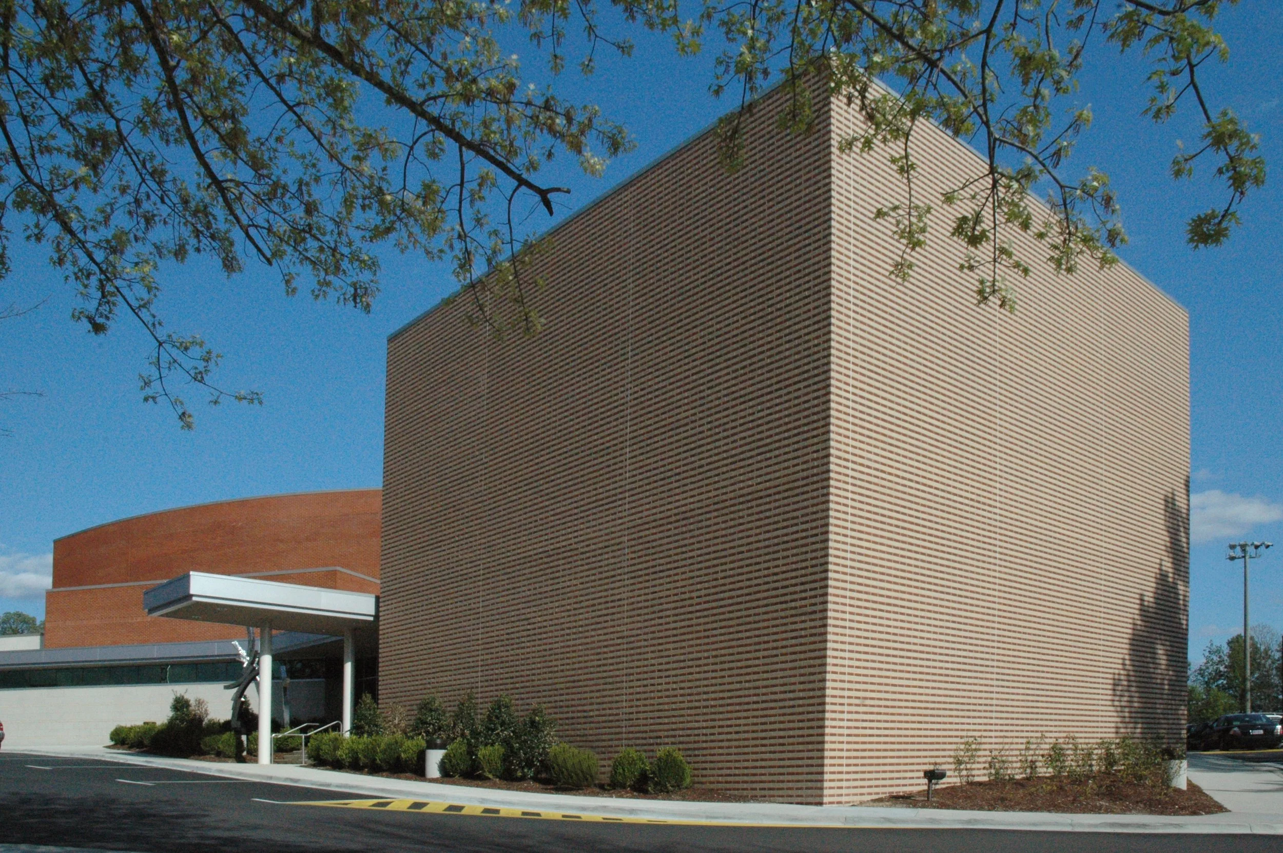 Modern brick building with a prominent large brick wall, surrounded by a parking lot, trees, and clear blue sky.
