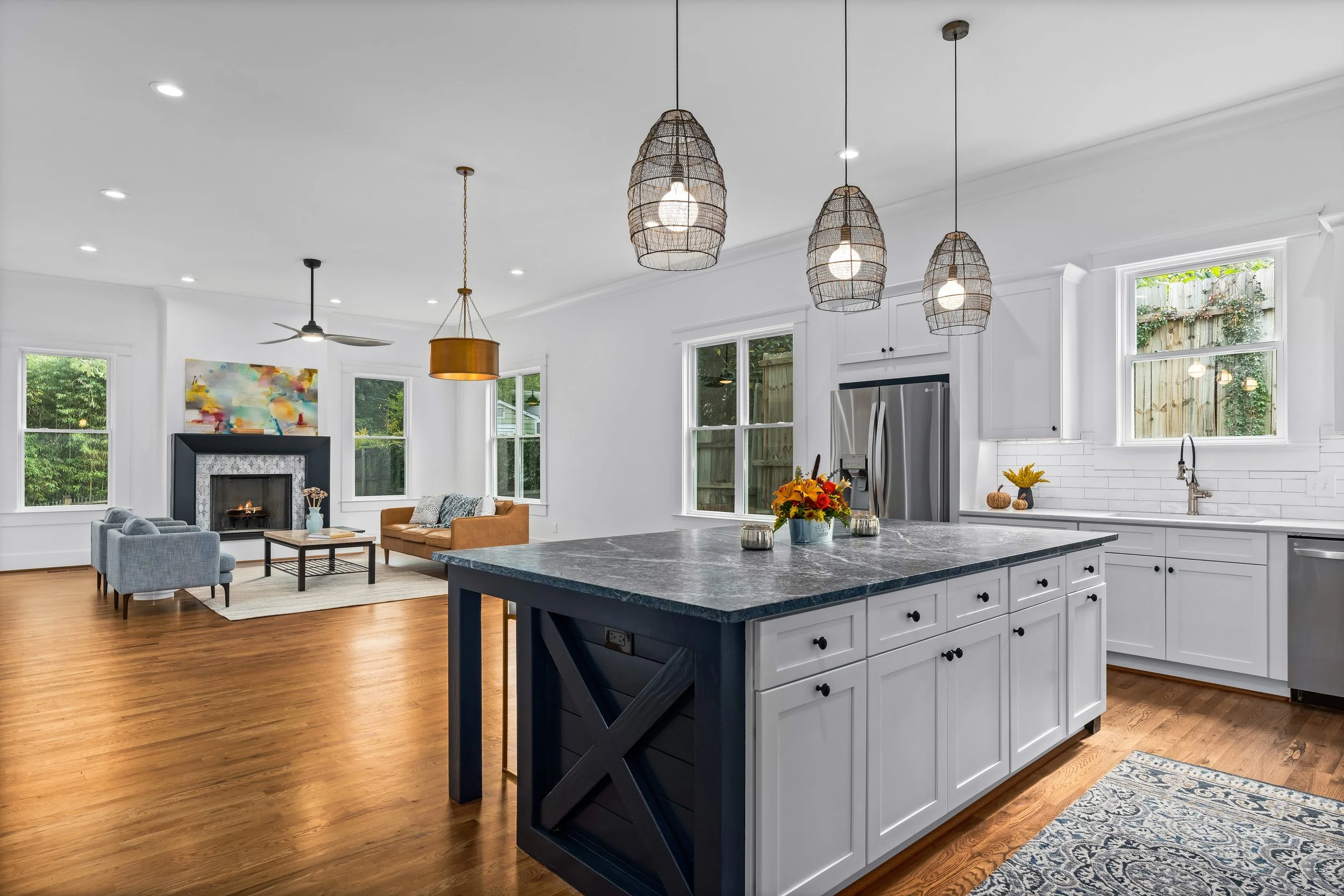 Open-concept living room and kitchen with white cabinets, stainless steel refrigerator, granite kitchen island, hardwood floors, three pendant lights, and a decorated fireplace.