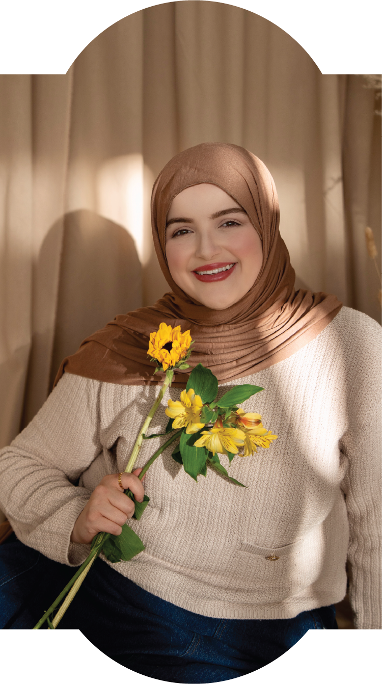 A woman with a brown hijab smiling and holding a bouquet of yellow flowers indoors.