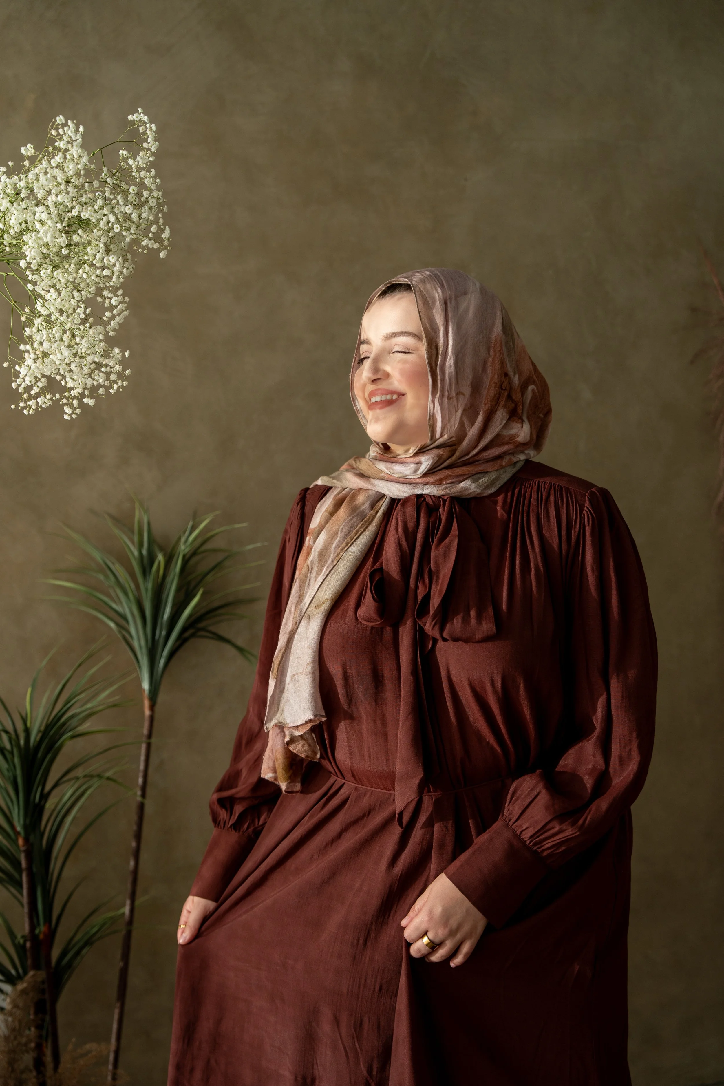 A smiling woman wearing a brown dress and a beige scarf stands in front of a greenish background with plants, looking happy.