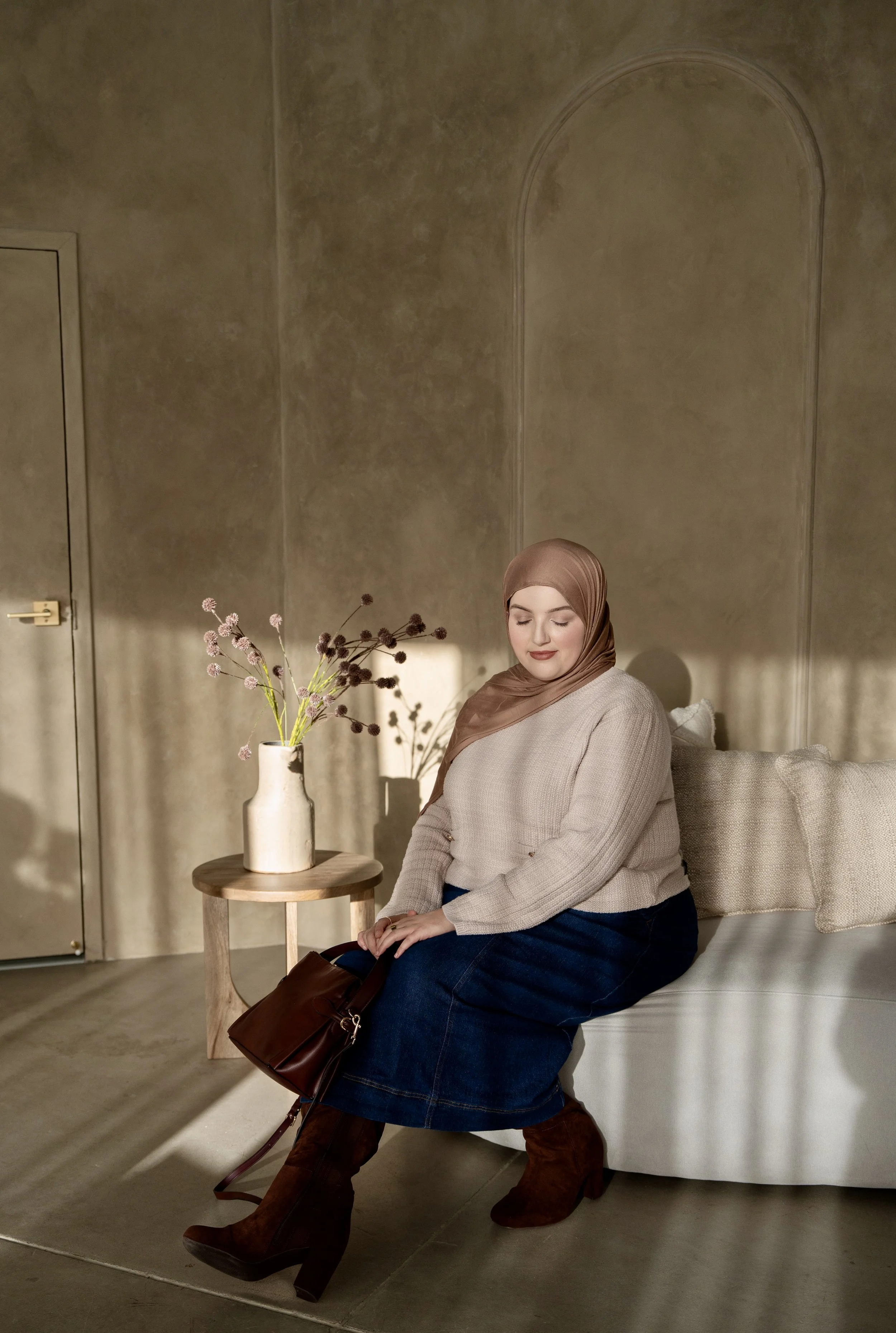 A woman wearing a brown hijab, light green blouse, and beige pants sitting on a modern white armchair in a room with beige curtains and a wooden floor. There is a cream-colored handbag with a gold logo on the floor next to her.