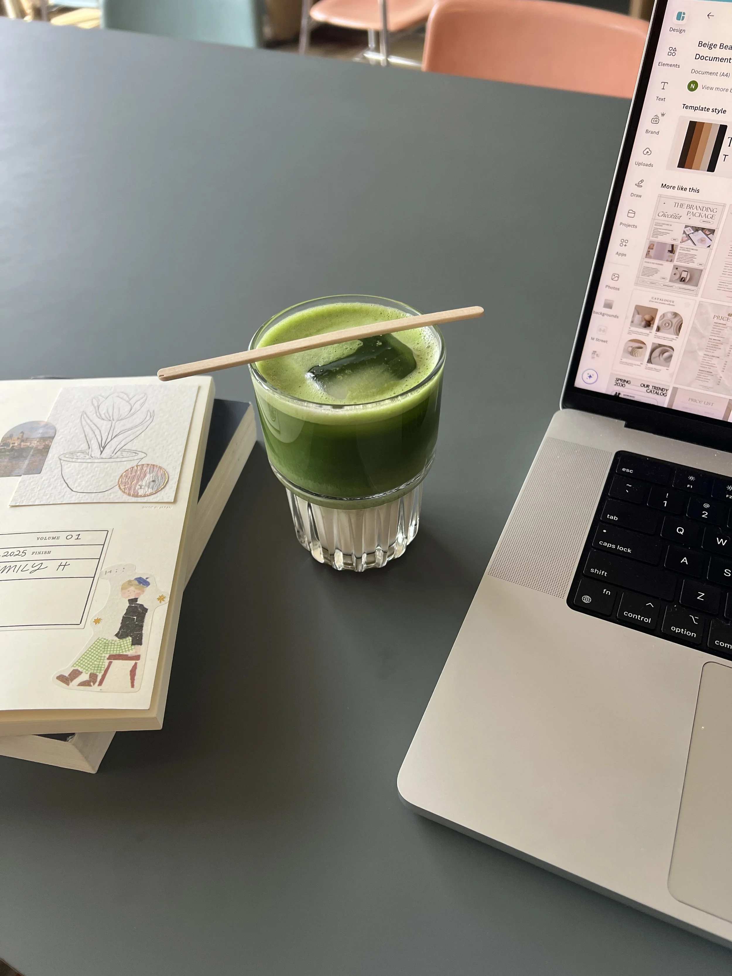 A desk with a book, a glass of green juice with a straw, and a laptop open to a webpage in a coffee shop or office setting.