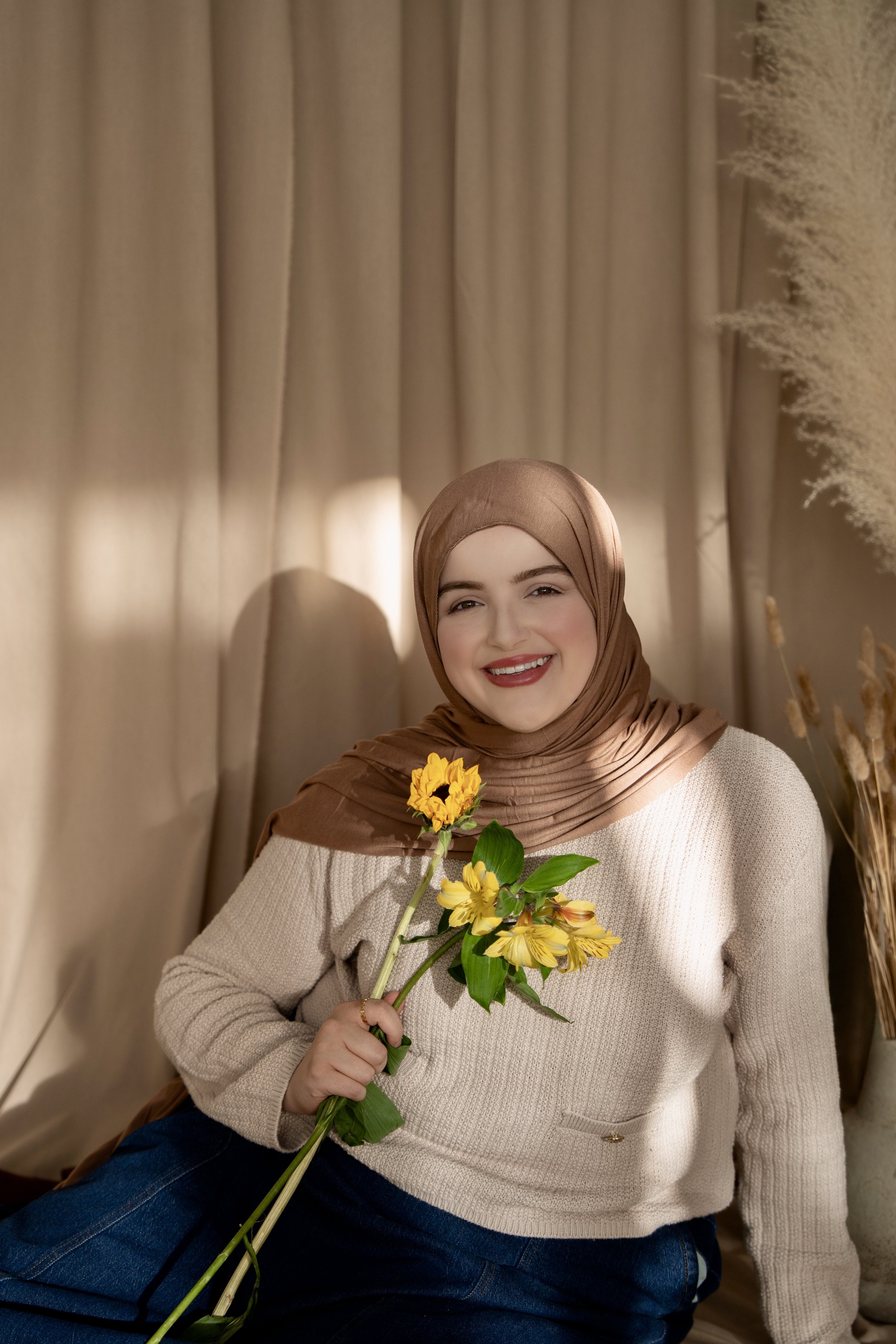 Muslim professional wearing a brown hijab and neutral sweater, smiling softly with yellow flowers in hand, photographed in a clean, minimalist studio.