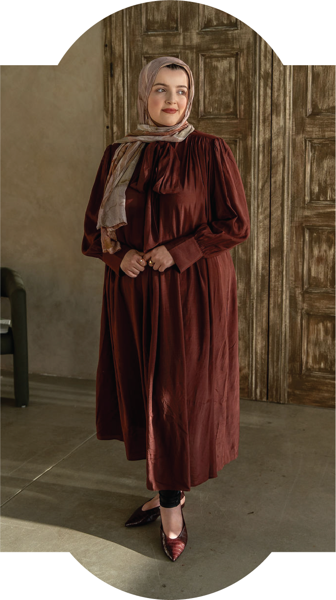A woman is standing indoors wearing a long dark red dress with a matching scarf, standing in front of wooden doors.