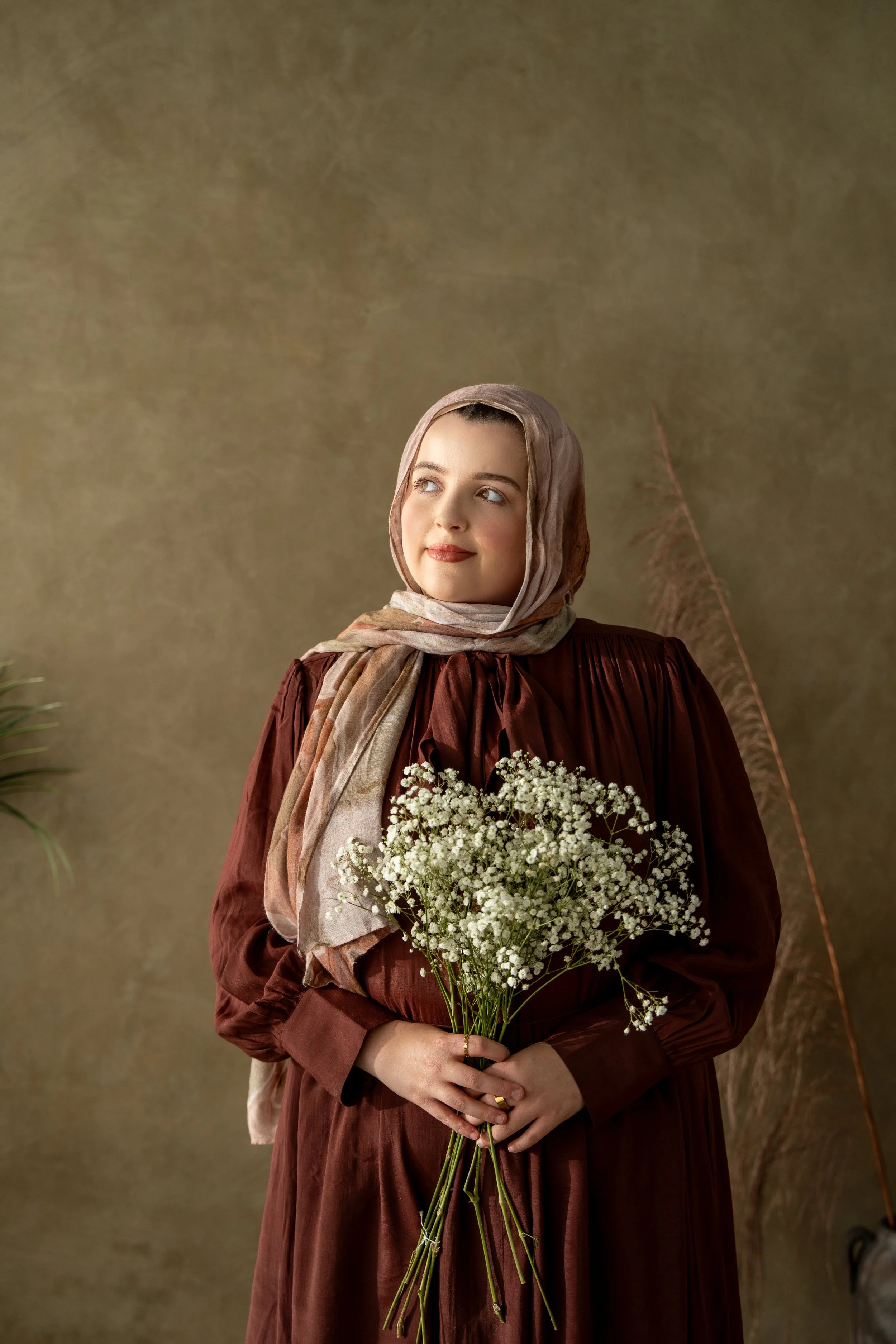 Plus-size Muslim woman wearing a beige hijab and burgundy dress, holding white flowers and looking upward in a serene, earthy-toned studio