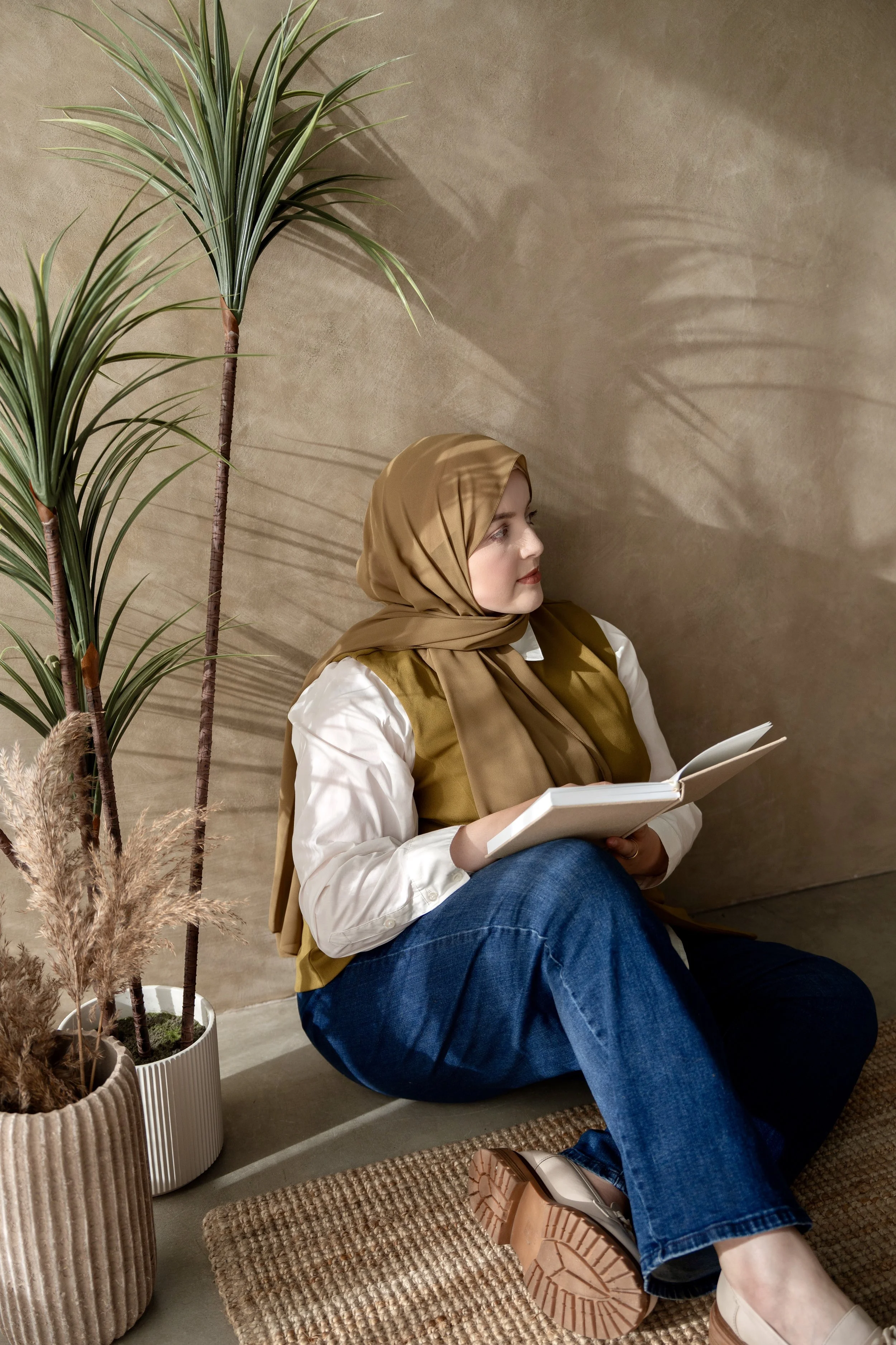A woman wearing a tan headscarf, white shirt, and blue jeans is sitting on the floor, reading a book. She is near a beige wall with two tall green potted plants next to her, and she is seated on a small woven rug.