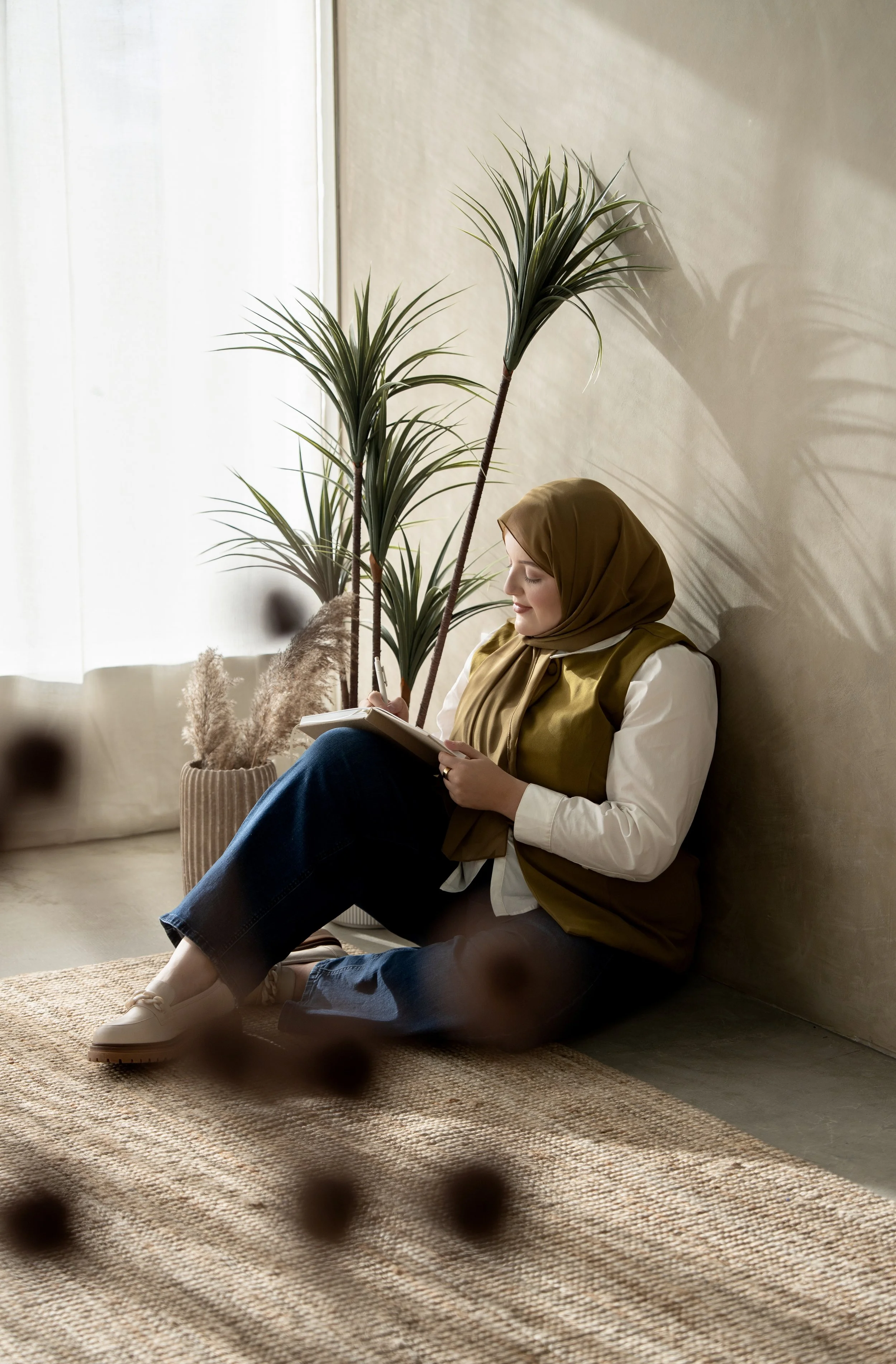 A woman wearing a brown hijab, white shirt, and olive vest sitting on a beige rug against a wall. She is reading a book, with tall indoor plants and a vase with pampas grass nearby.