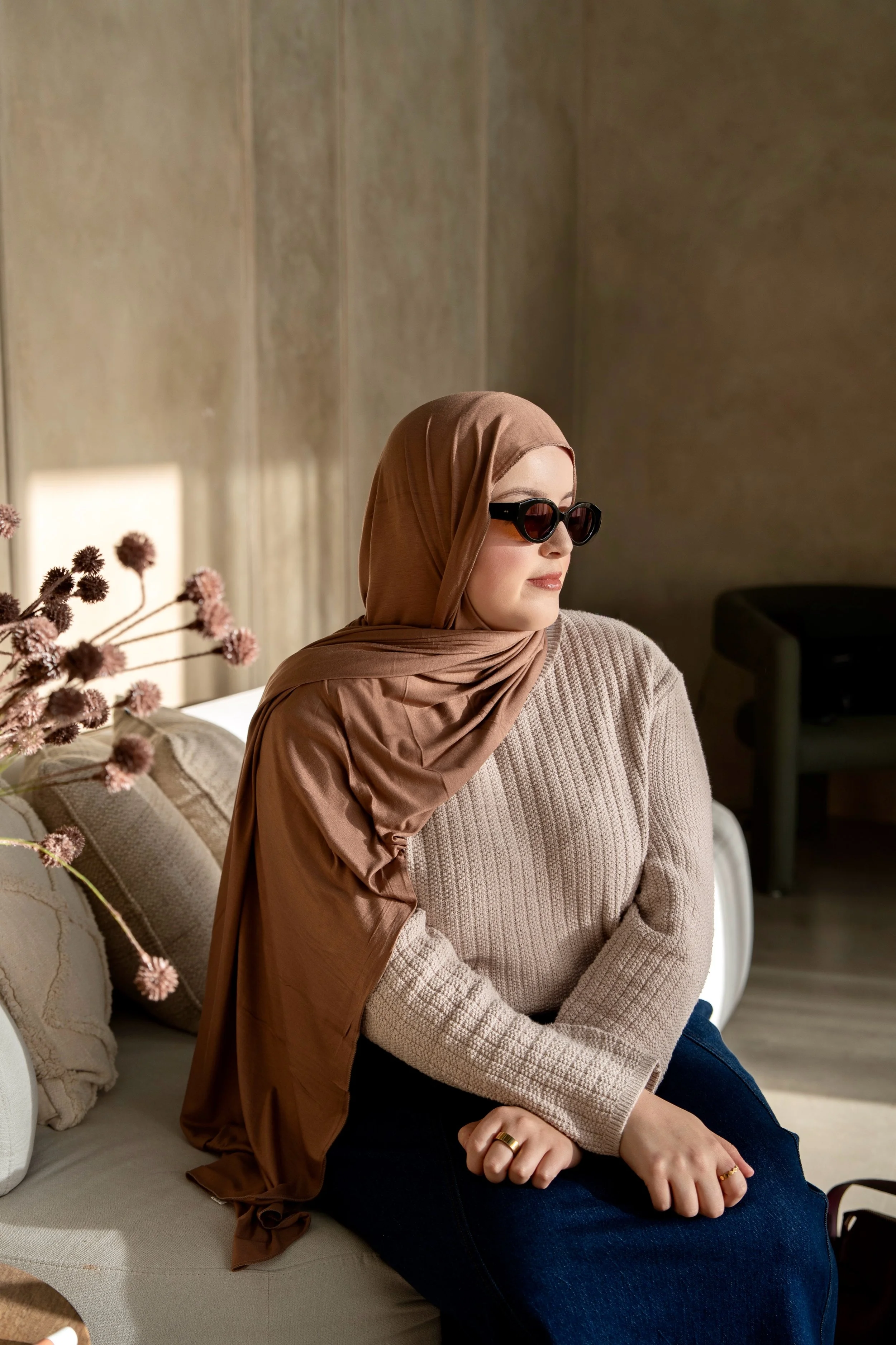 A woman wearing a brown hijab and black sunglasses sitting on a beige sofa with a beige knitted sweater and blue jeans, with a background of beige walls and some decorative pillows.