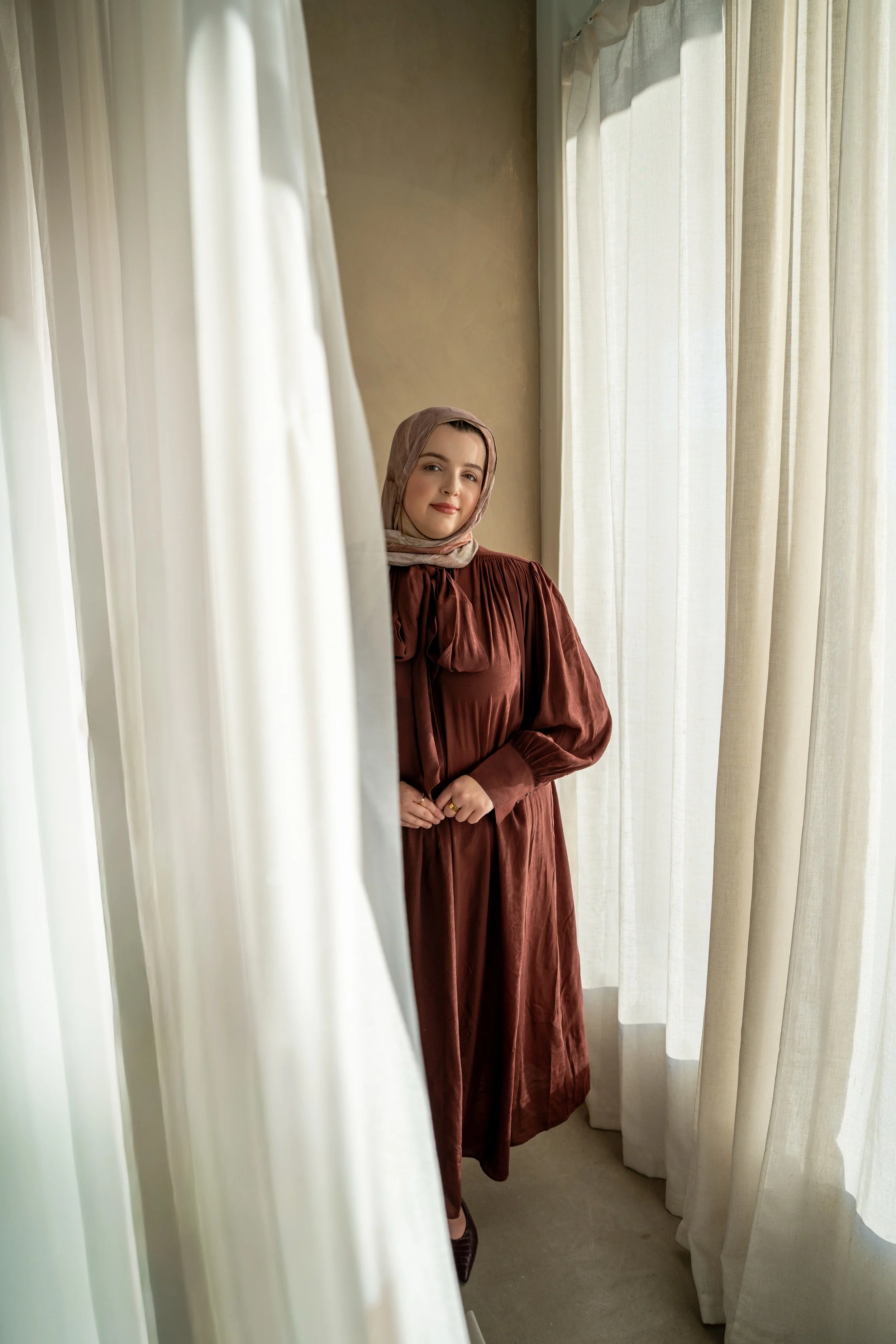A woman standing by a window, looking at the camera, wearing a brown dress and a light-colored headscarf, with white curtains around her.