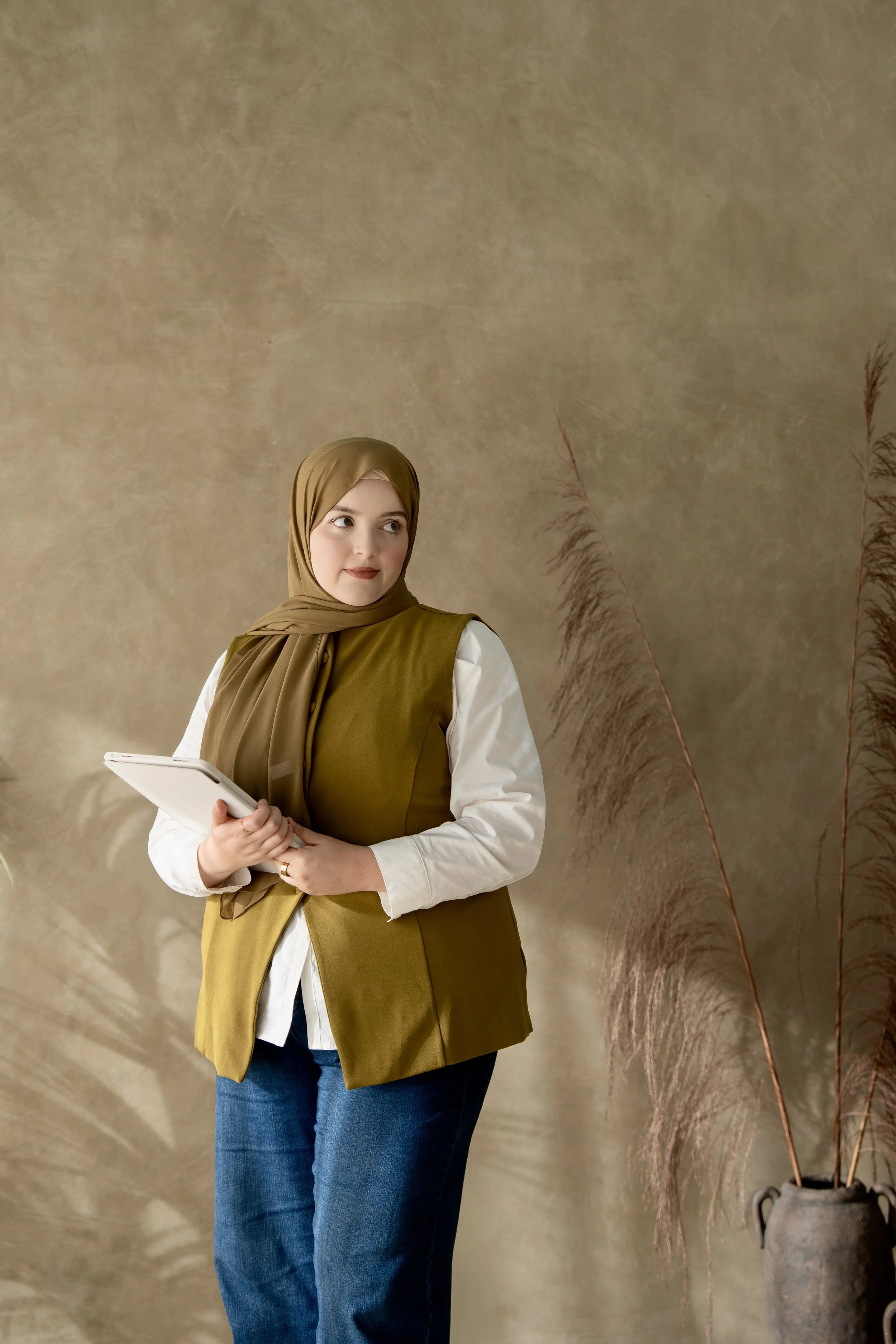 Muslim woman in an olive hijab and matching vest holding a tablet in a neutral studio space with natural décor.
