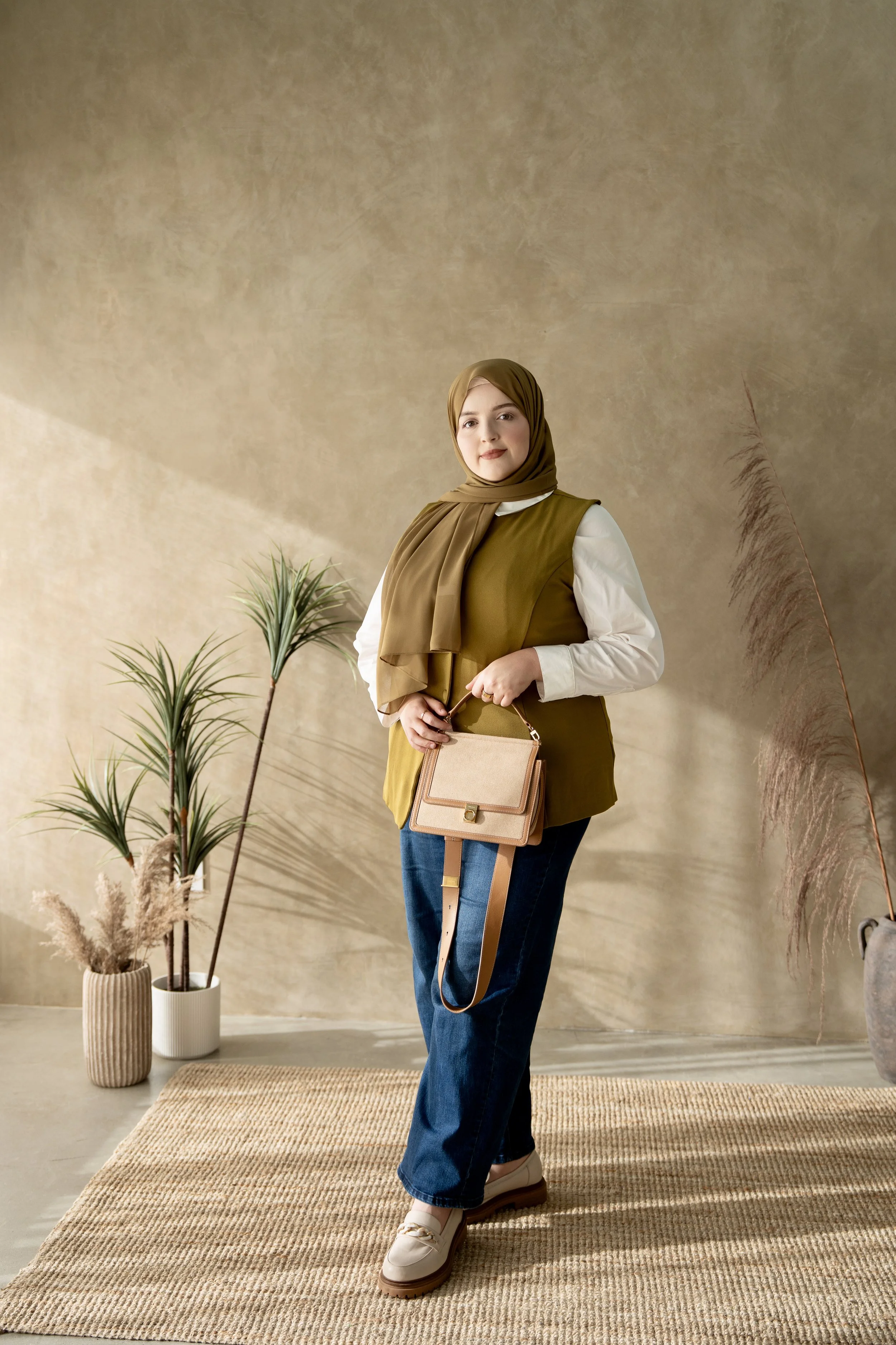 A woman wearing a brown hijab, white shirt, and olive green vest, holding a beige handbag, standing on a beige rug in a room with a neutral wall and potted plants.
