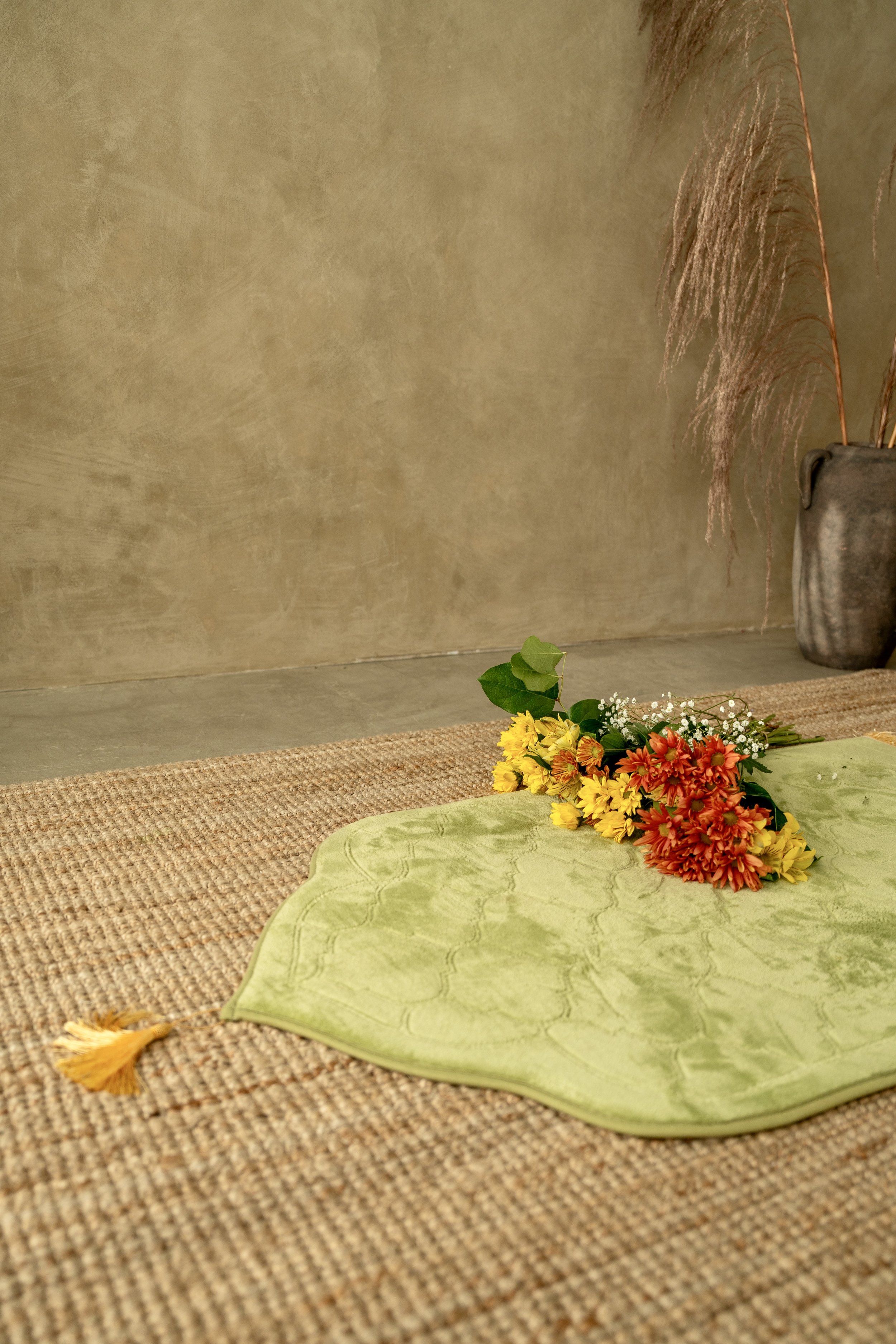 A bouquet of yellow and orange flowers with green leaves placed on a light green cloth, on a woven rug, with dried ornamental grasses in a vase in the background.
