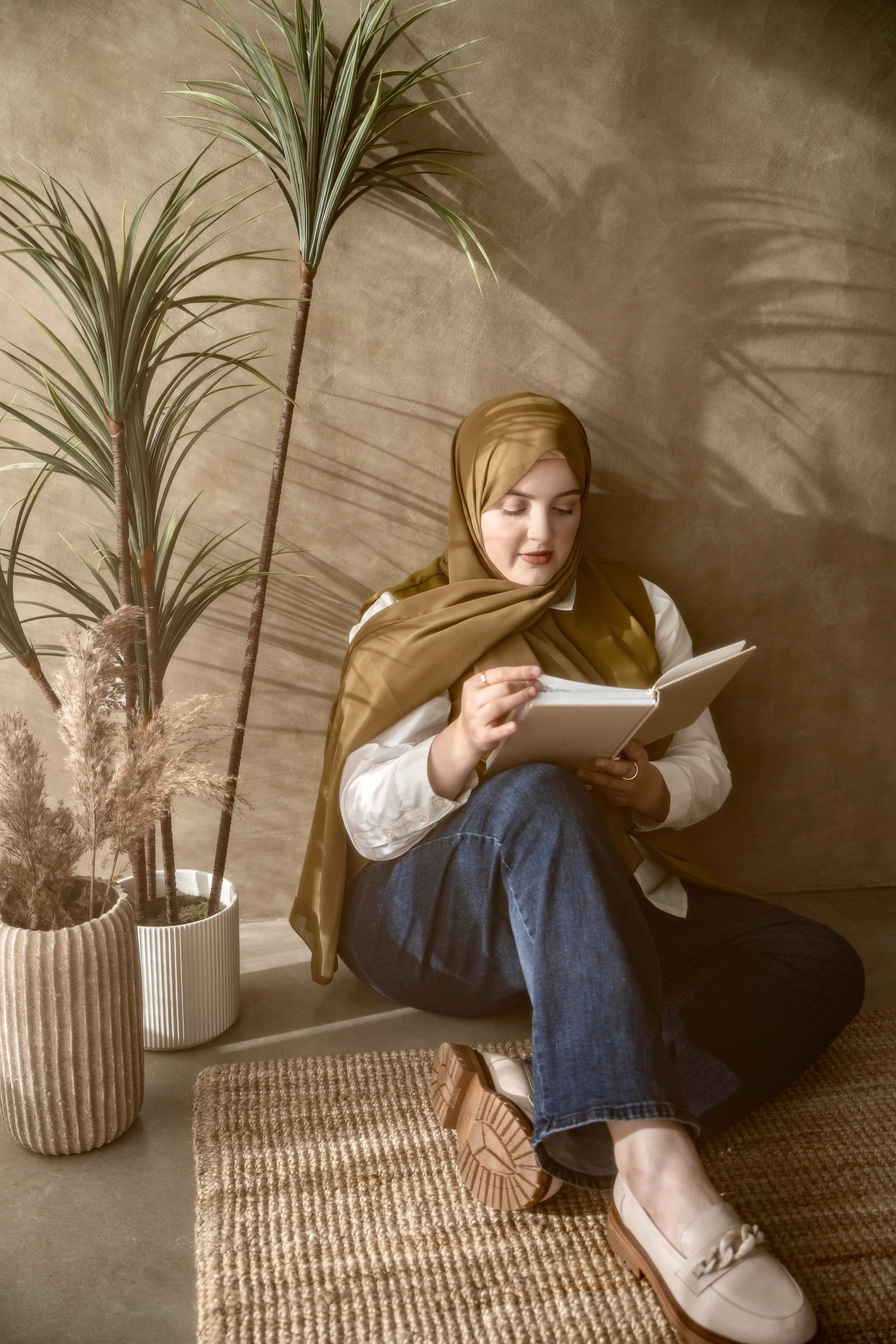 A woman wearing a brown hijab, white shirt, and blue jeans sits on a carpeted floor reading a book near two potted plants against a beige wall.