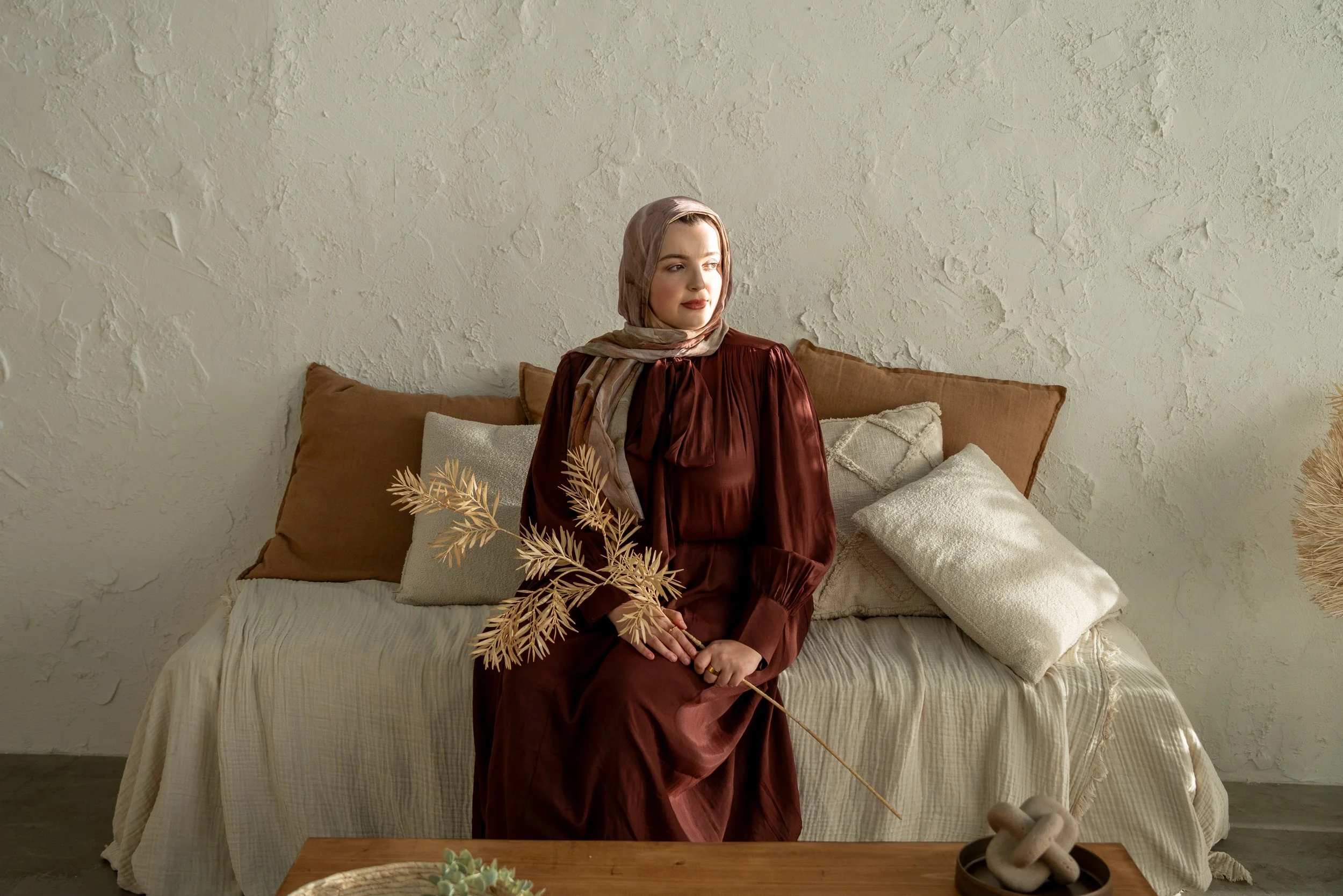 A woman in a brown dress and a beige headscarf sits on a bed with beige and brown pillows, holding a dried plant. The room has textured white walls and a wooden table with decorative items in the foreground.
