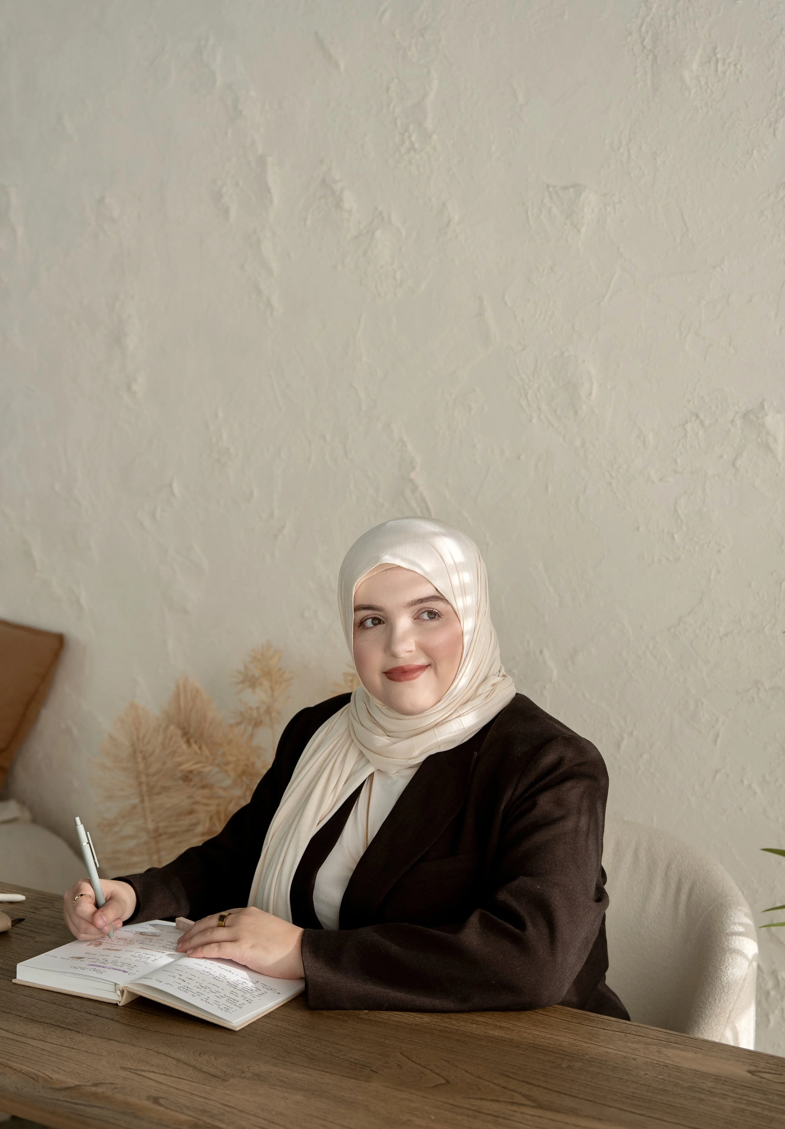 Empowering Muslim woman coach writing in her journal during a creative planning session, styled in a cream hijab and brown blazer with soft natural light and minimalist décor