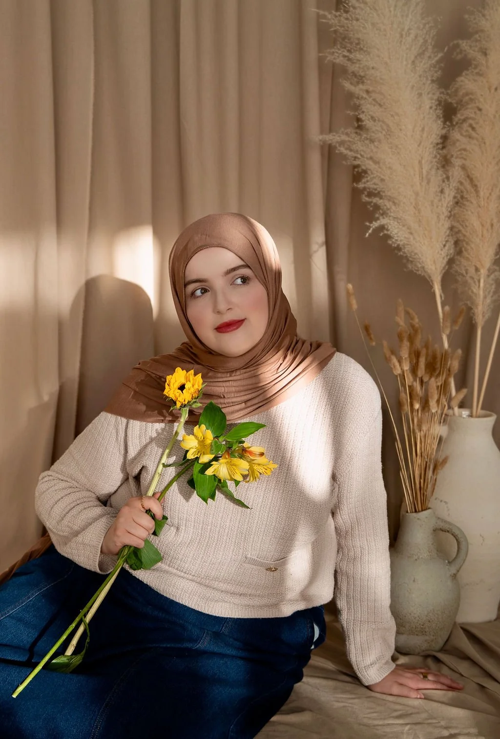 A woman in a beige sweater and brown headscarf sitting indoors with beige curtains in the background. She has light makeup, red lipstick, and is holding a small bouquet of yellow flowers. There is a large white vase with dried plants beside her.