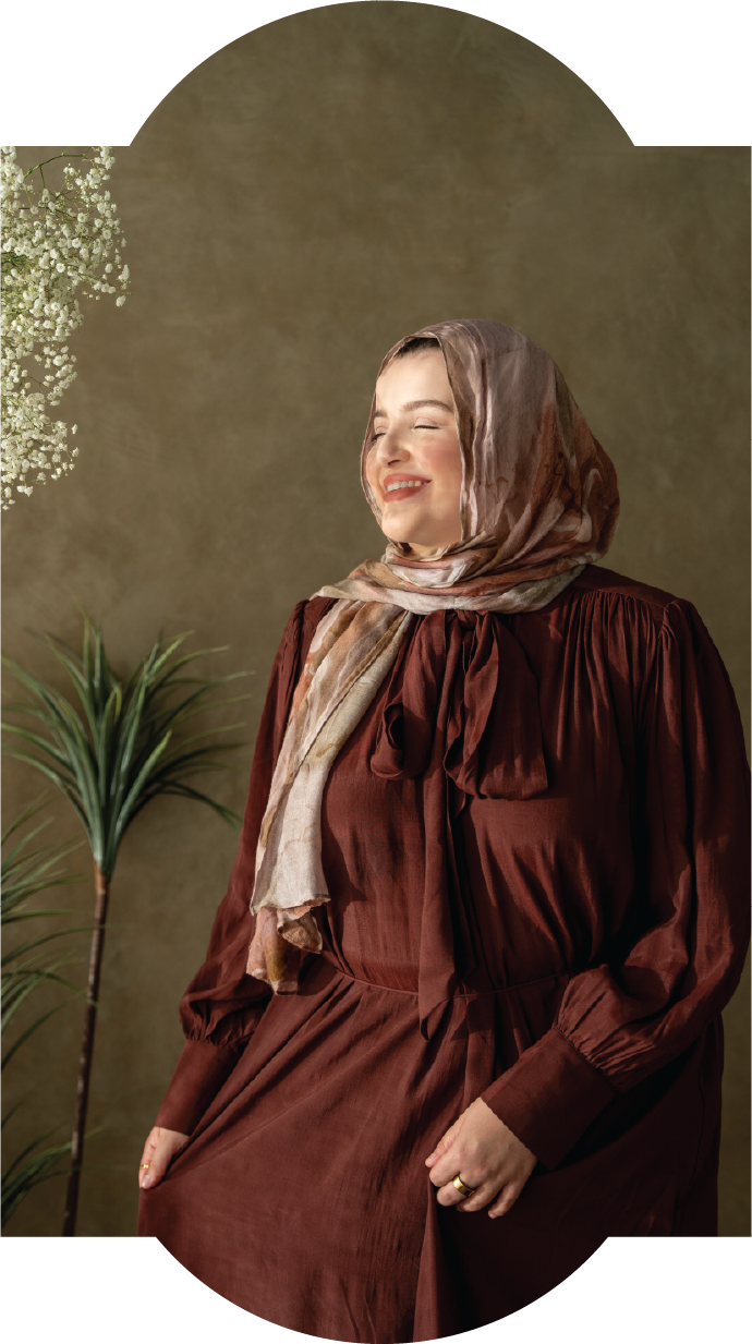 Woman smiling with eyes closed, wearing a brown dress and a light patterned scarf, standing near a plant and flowers against a plain background.