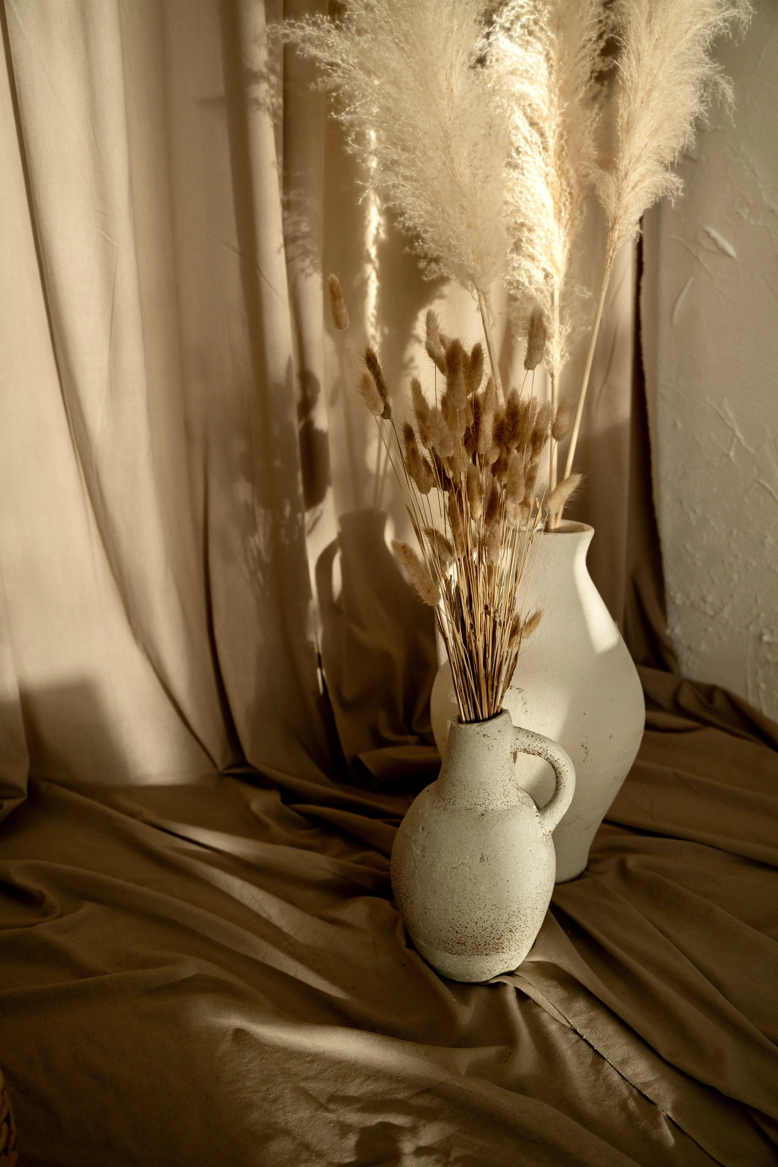Two white ceramic vases filled with dried pampas grass and other dried plants, set against beige curtains and velvet fabric.