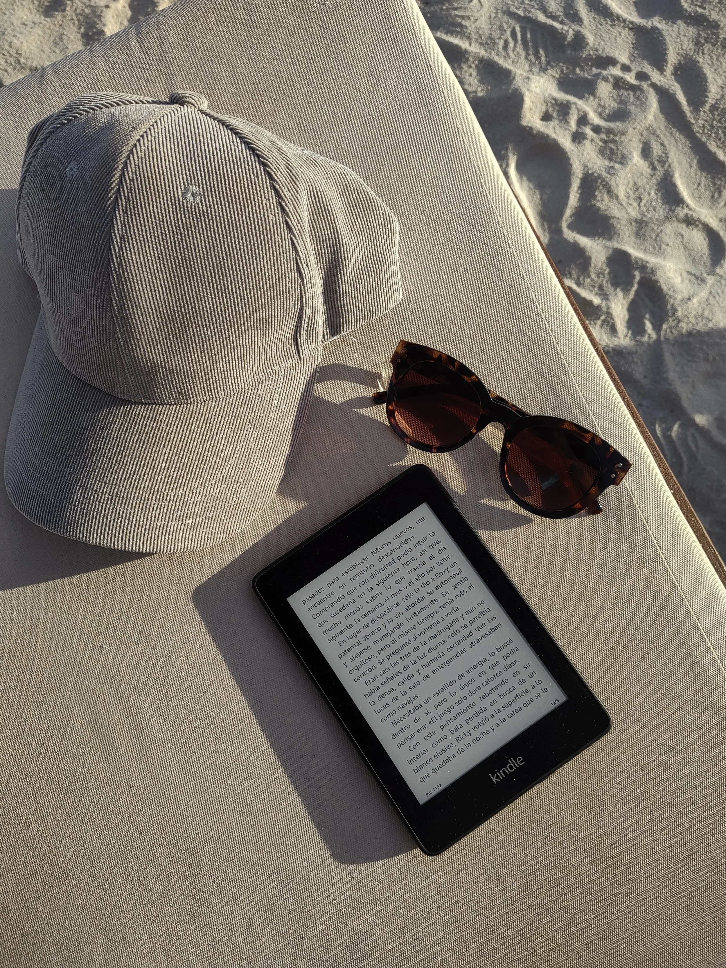A beige baseball cap, tortoiseshell sunglasses, and a Kindle e-reader on a cream-colored lounge chair at the beach.