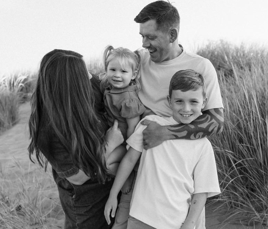 A happy family of four on the beach, smiling and laughing. The father is holding a young girl, with a teenage boy standing beside him, and a woman, likely the mother, is also present.
