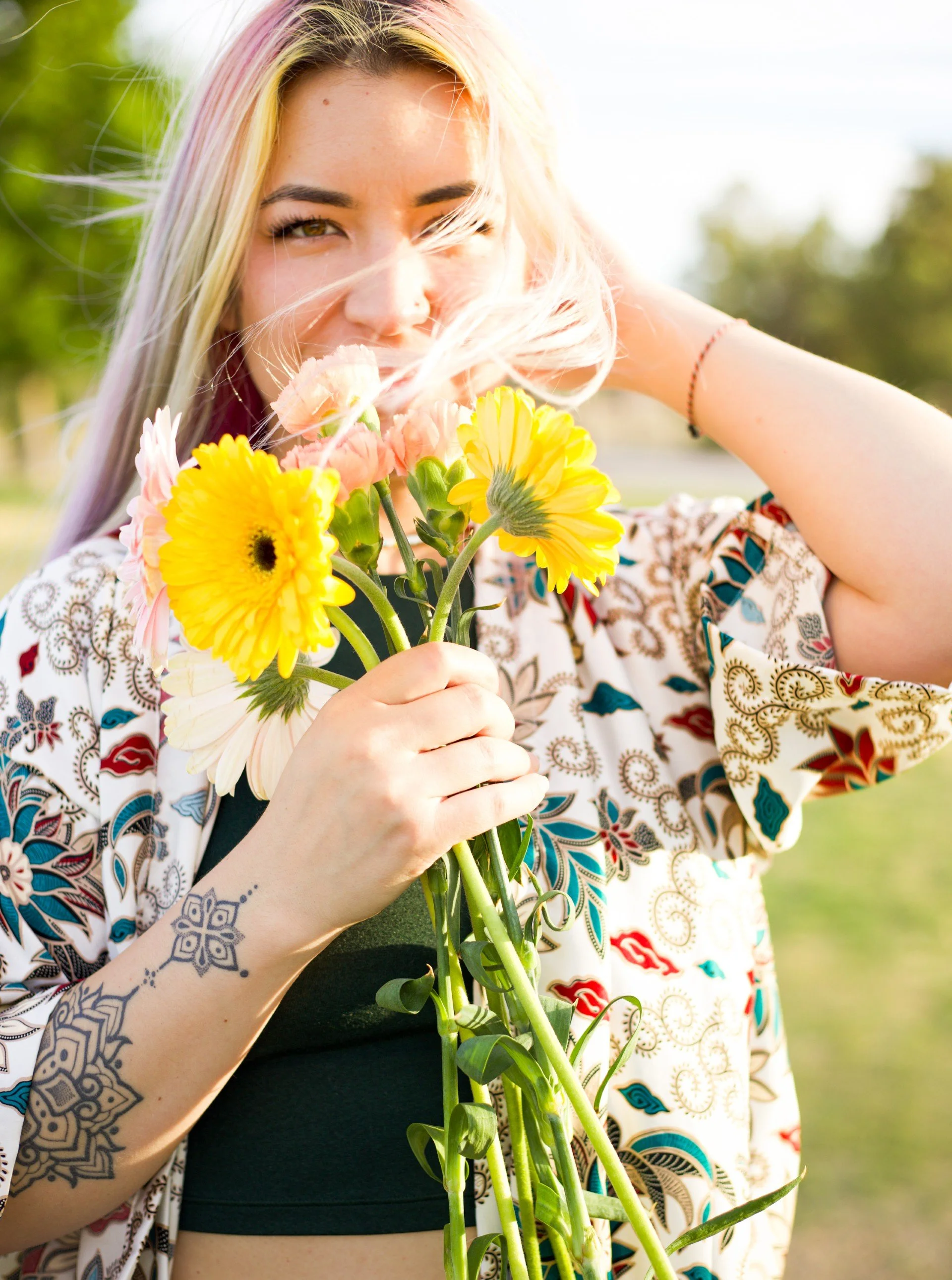 A woman with blonde hair holding a bouquet of mixed yellow and pink flowers, with a tattoo on her arm, standing outdoors on a sunny day.
