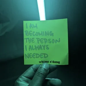 A green card with the handwritten message 'I am becoming the person I always needed' and the words 'whims o'dang' at the bottom, held by a gloved hand under a teal light.