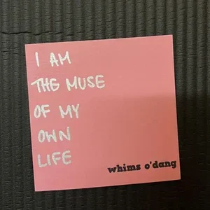 A pink sticky note with white handwritten text that says 'I am the muse of my own life' and a black printed phrase 'whims o'dang'.