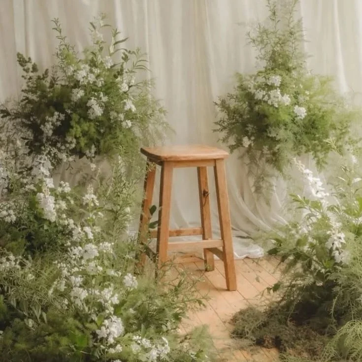 A wooden stool surrounded by white flowering plants with a white curtain backdrop.
