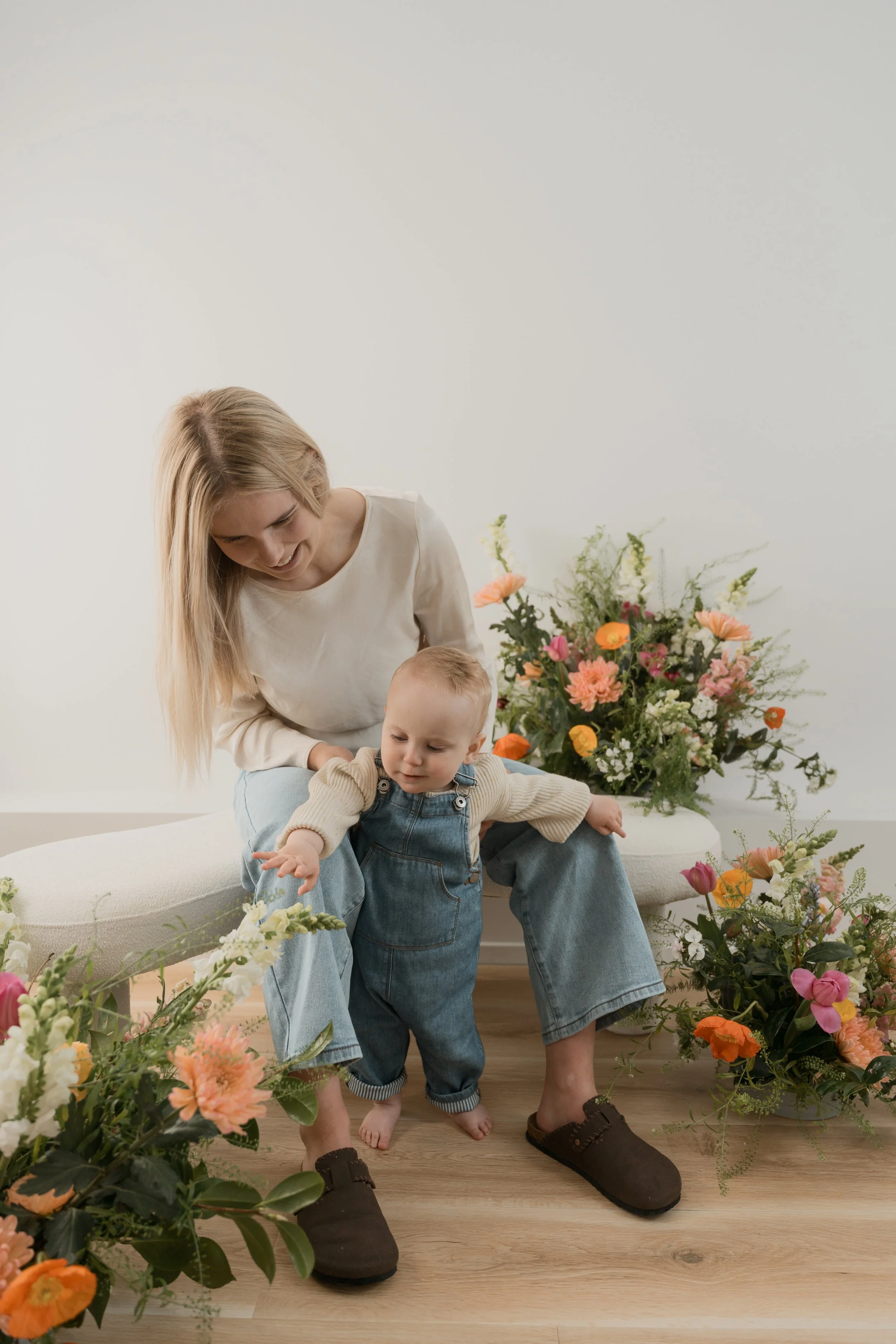 A woman and two young children sitting on a bench surrounded by numerous colorful flowers and greenery, in a bright, minimalistic space.