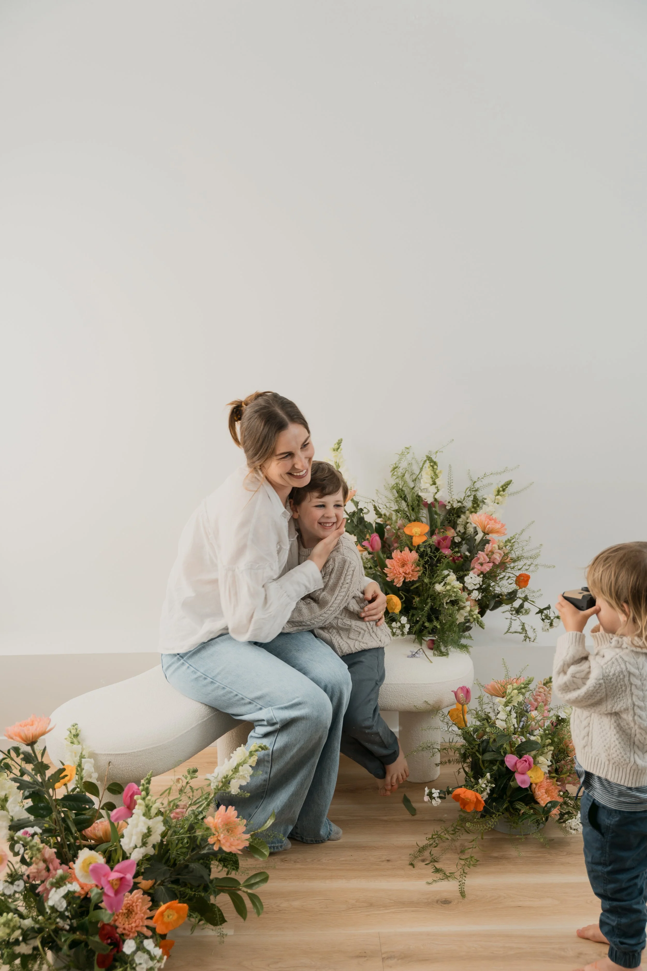 A woman and a young girl are hugging each other and smiling near a large flower arrangement. There are other children and more flowers in the scene, with a plain white wall in the background.