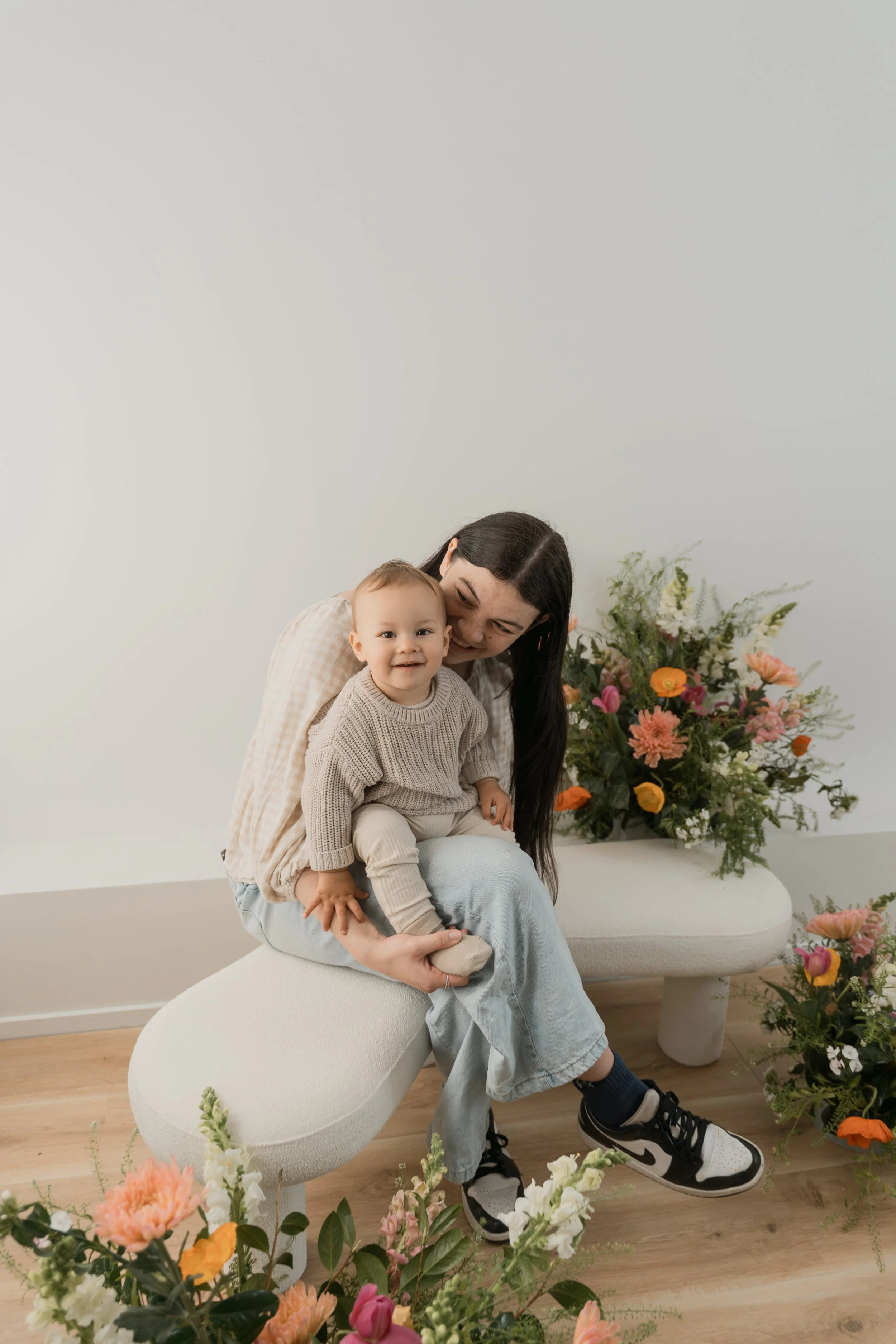 A woman with long dark hair and a young boy with short light hair sitting on a white modern bench surrounded by a variety of colorful flowers, set against a plain light-colored wall.