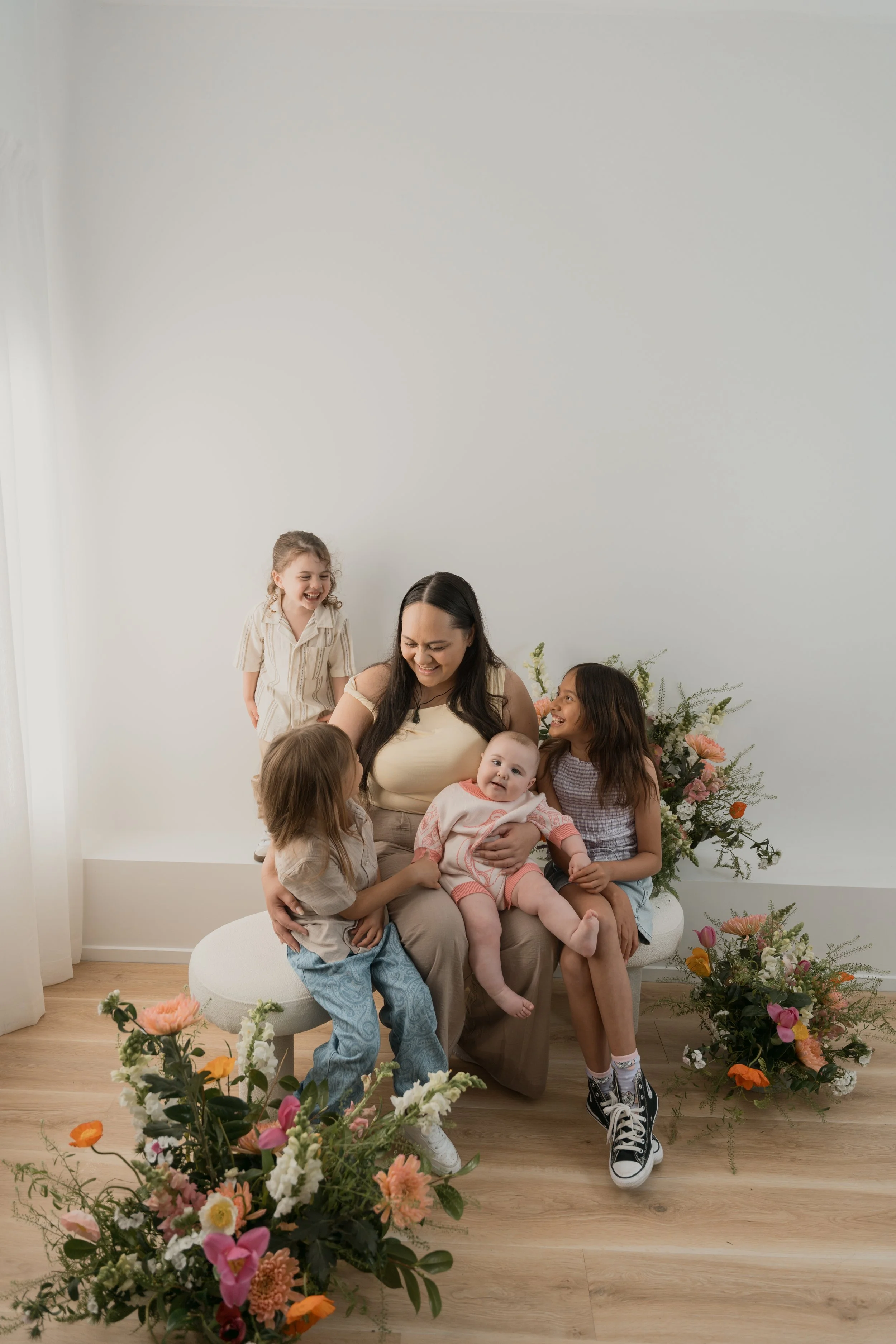 A group of children and a woman sitting on a white round ottoman in front of a plain white wall surrounded by large floral arrangements, with one girl standing behind them laughing.