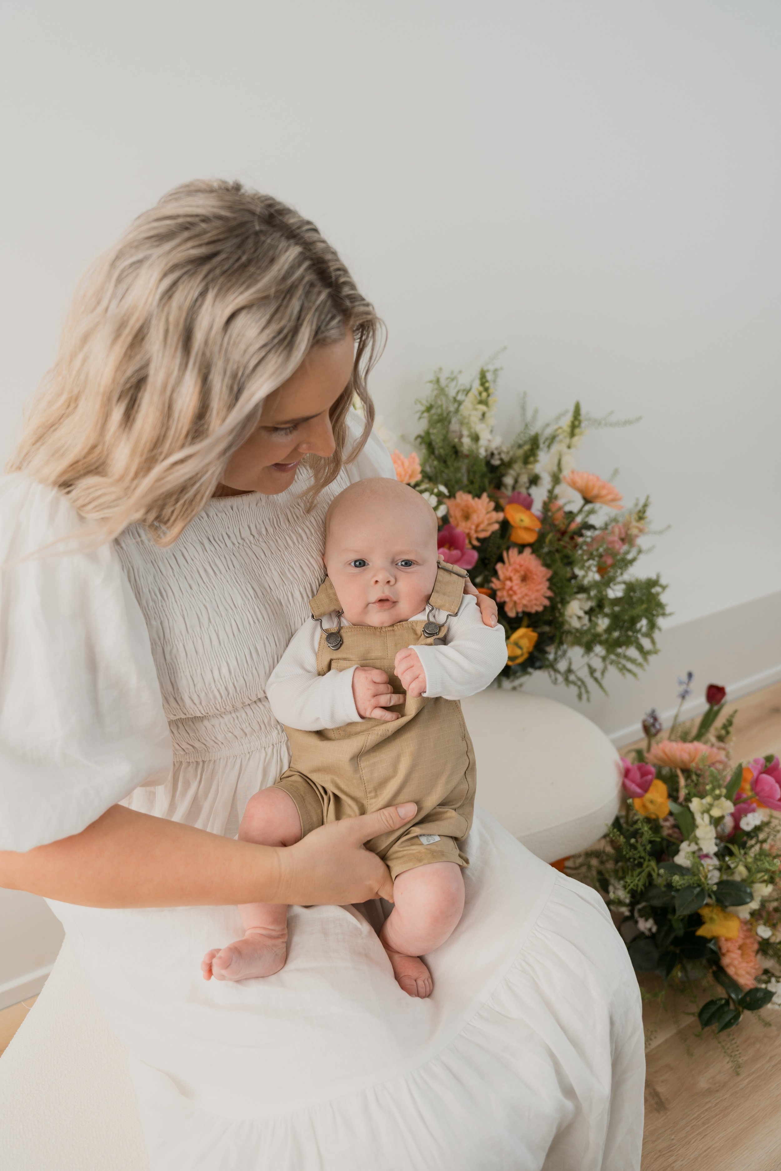 A woman with blonde hair holding a baby in a beige outfit, seated on a white cushioned chair with floral arrangements in the background.