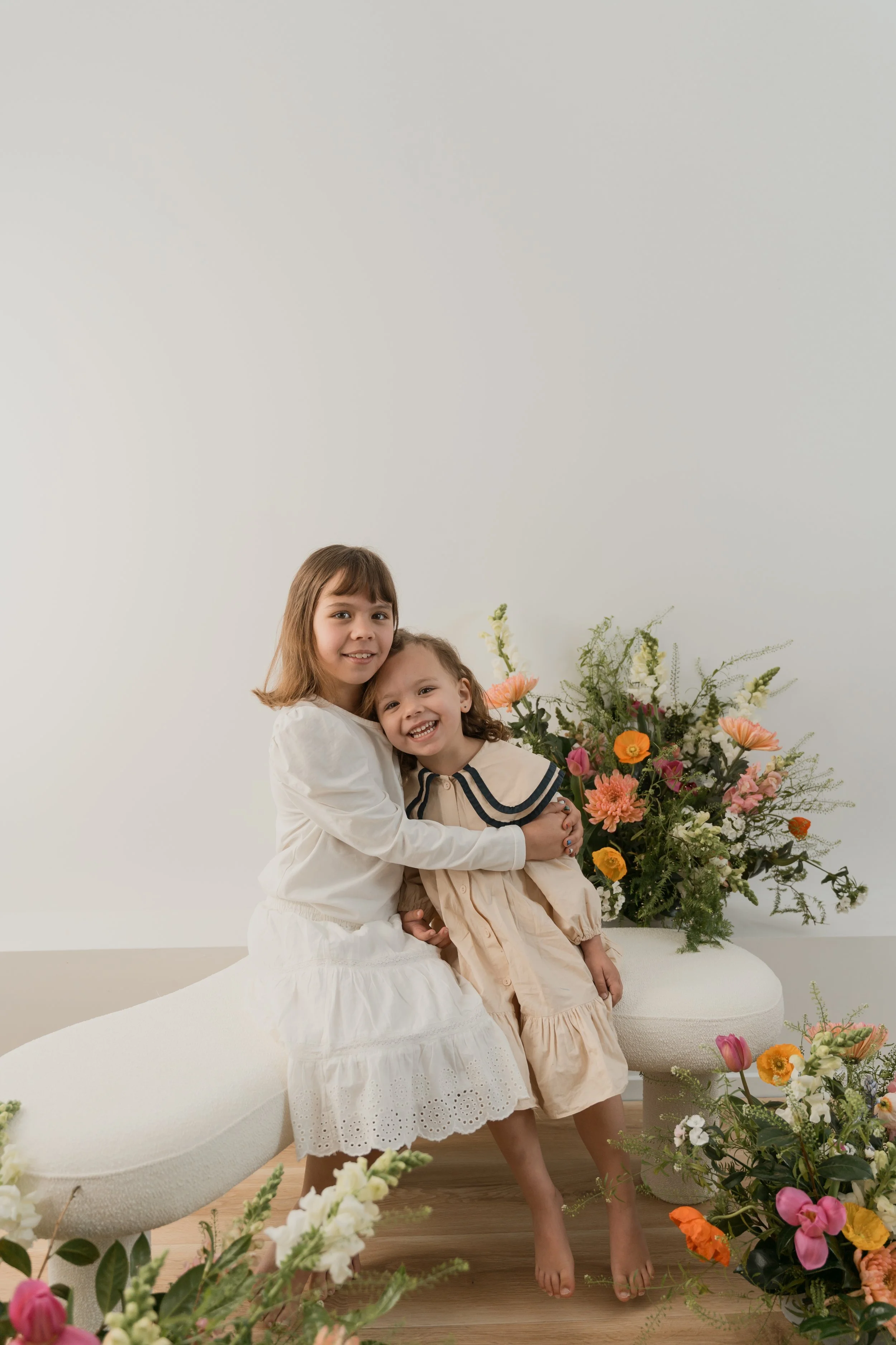 Two young girls sitting and standing next to each other on a white bench surrounded by colorful flowers, in a minimalistic, bright room.