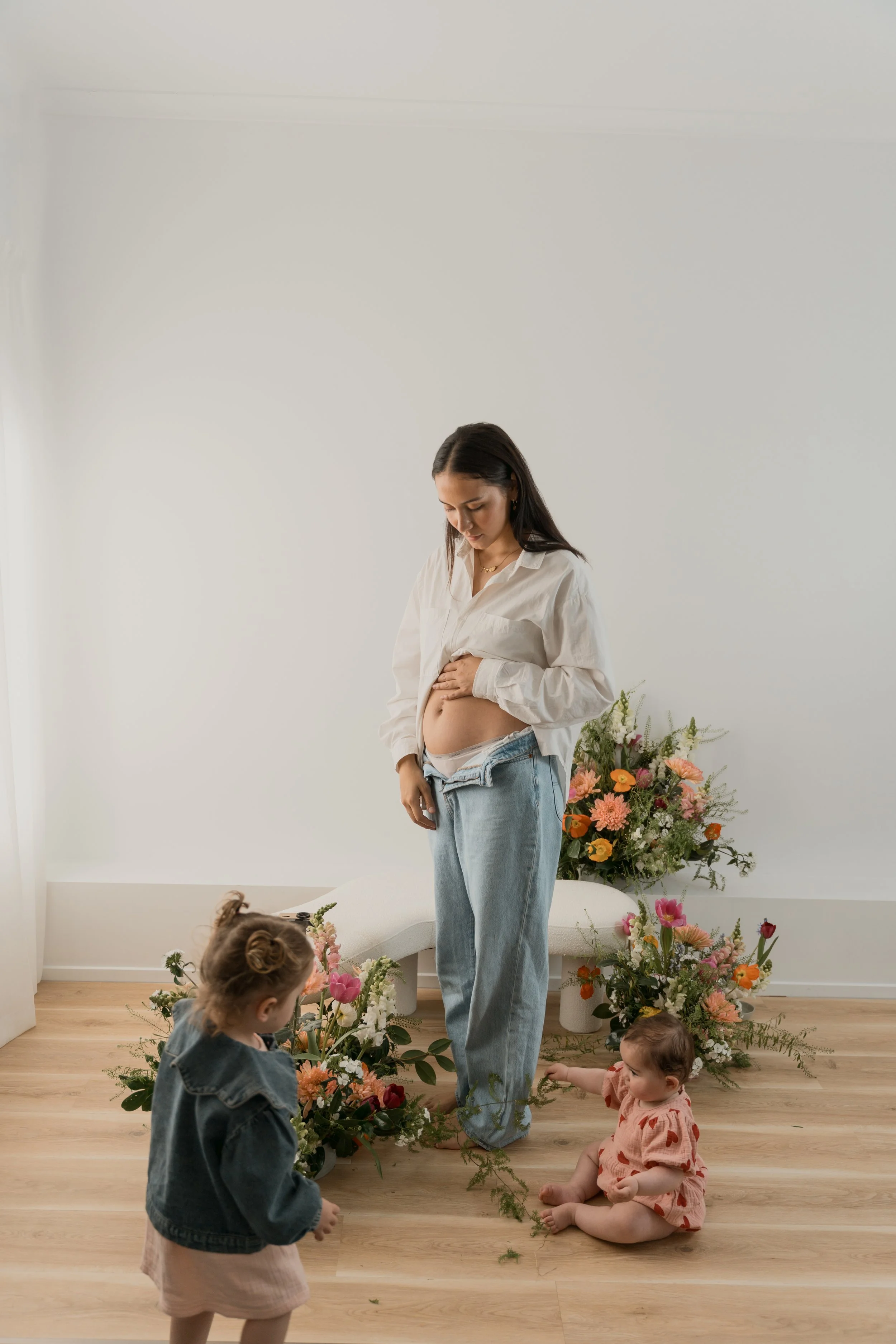 A woman with long dark hair, wearing a white shirt and blue jeans, standing in a room with a plain white wall, surrounded by children and flowers on the floor and in pots. The children are sitting and standing, engaging with flowers and greenery.