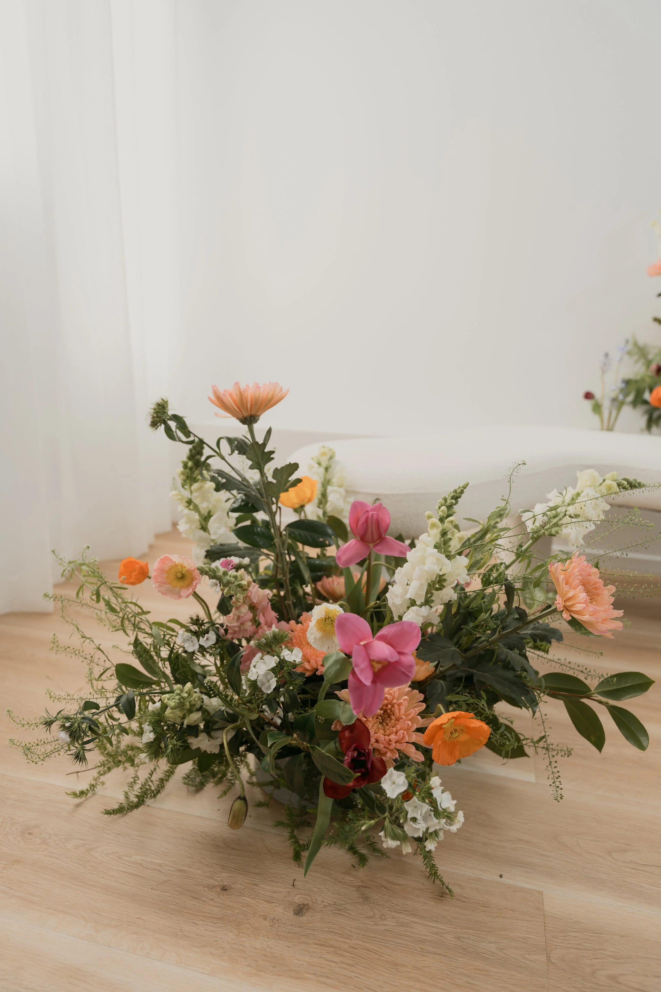 A vibrant bouquet of mixed flowers, including pink, orange, and white blooms, arranged with green foliage, placed on a light wooden floor against a minimalist white background.