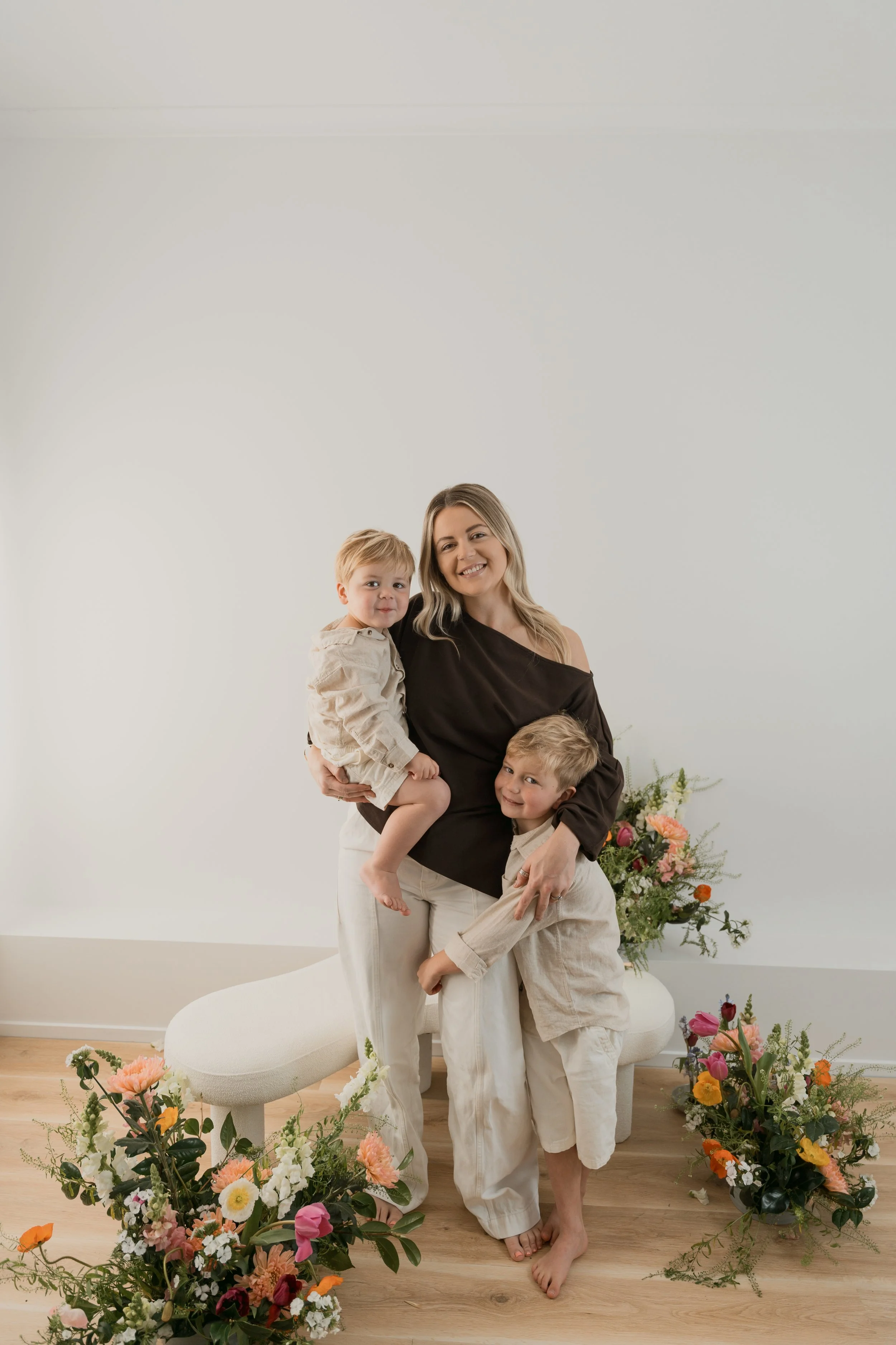 A woman smiling and holding two young boys, surrounded by colorful flowers in a bright, minimalist room.